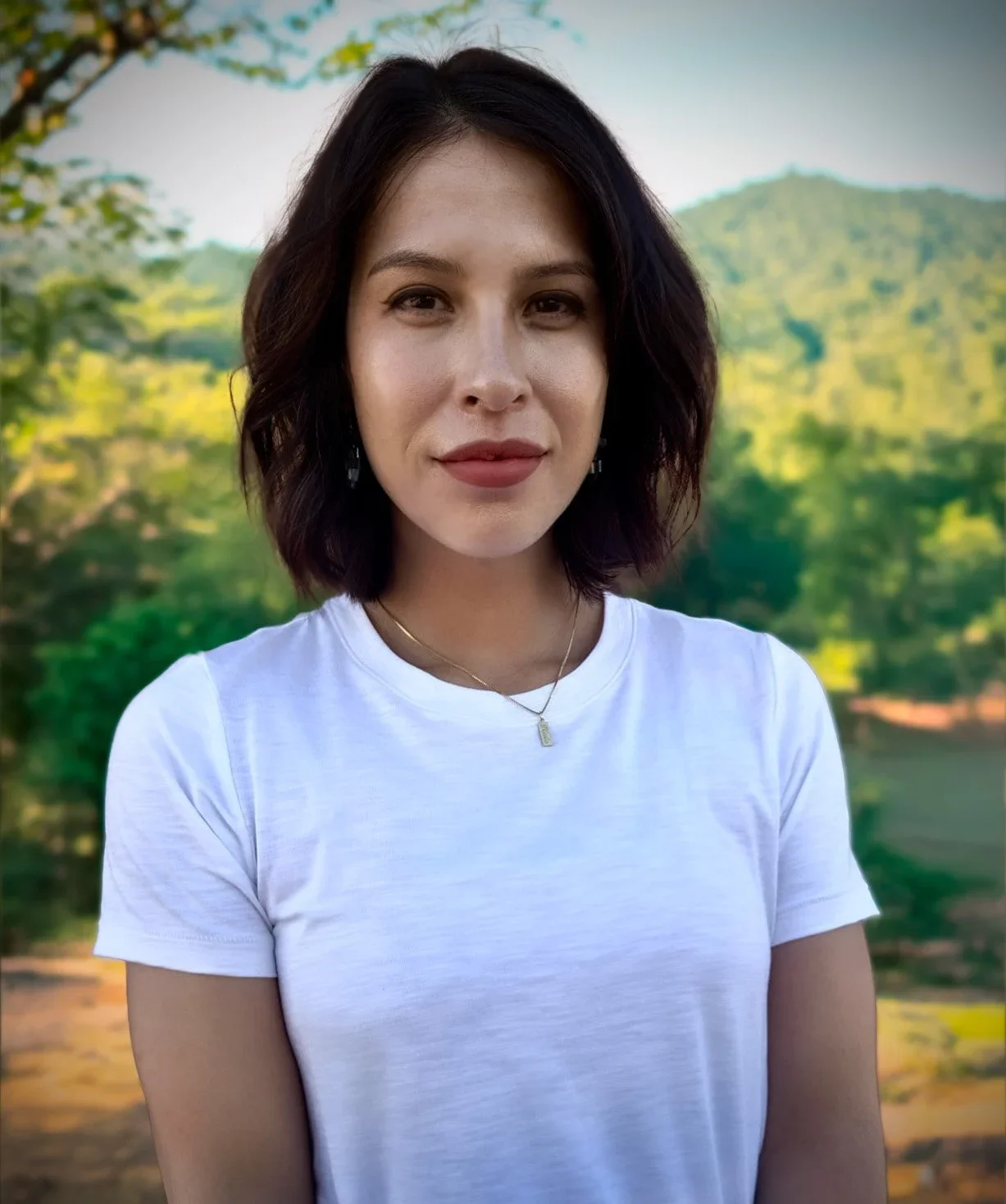 A woman with shoulder-length dark hair wearing a white t-shirt, standing outdoors with a background of green trees and hills.