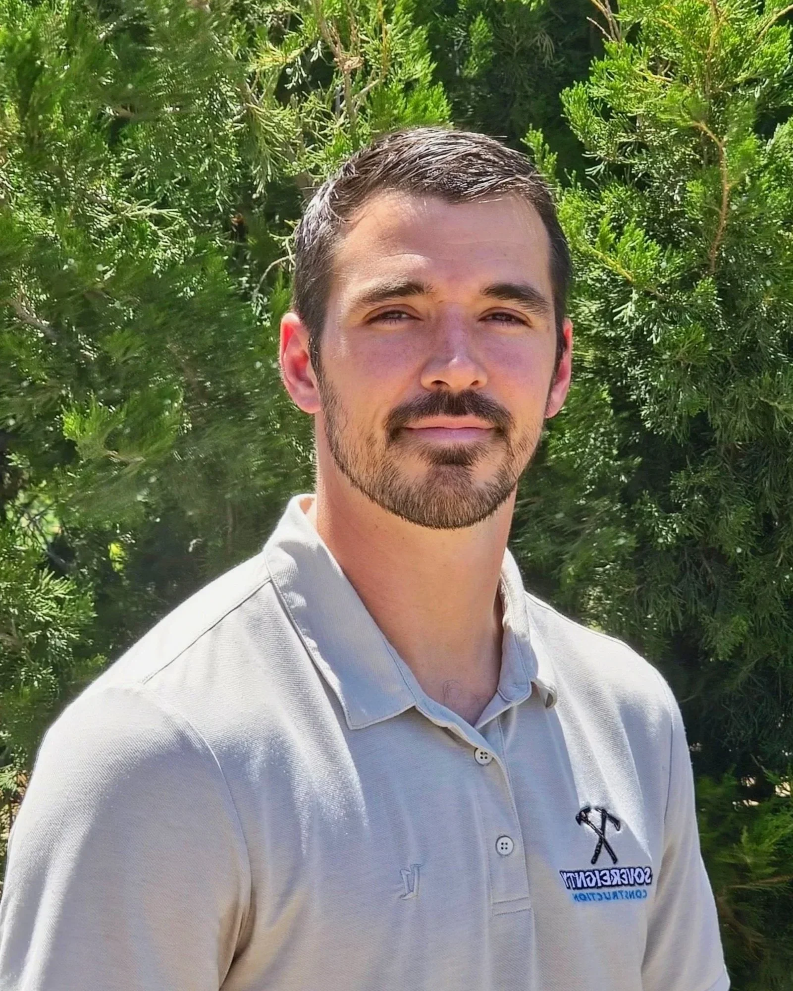 A young man with dark hair, a trimmed beard, and light skin posing outdoors in front of green trees, wearing a light-colored polo shirt with a logo that features a hammer and the word 'SERVCON'.
