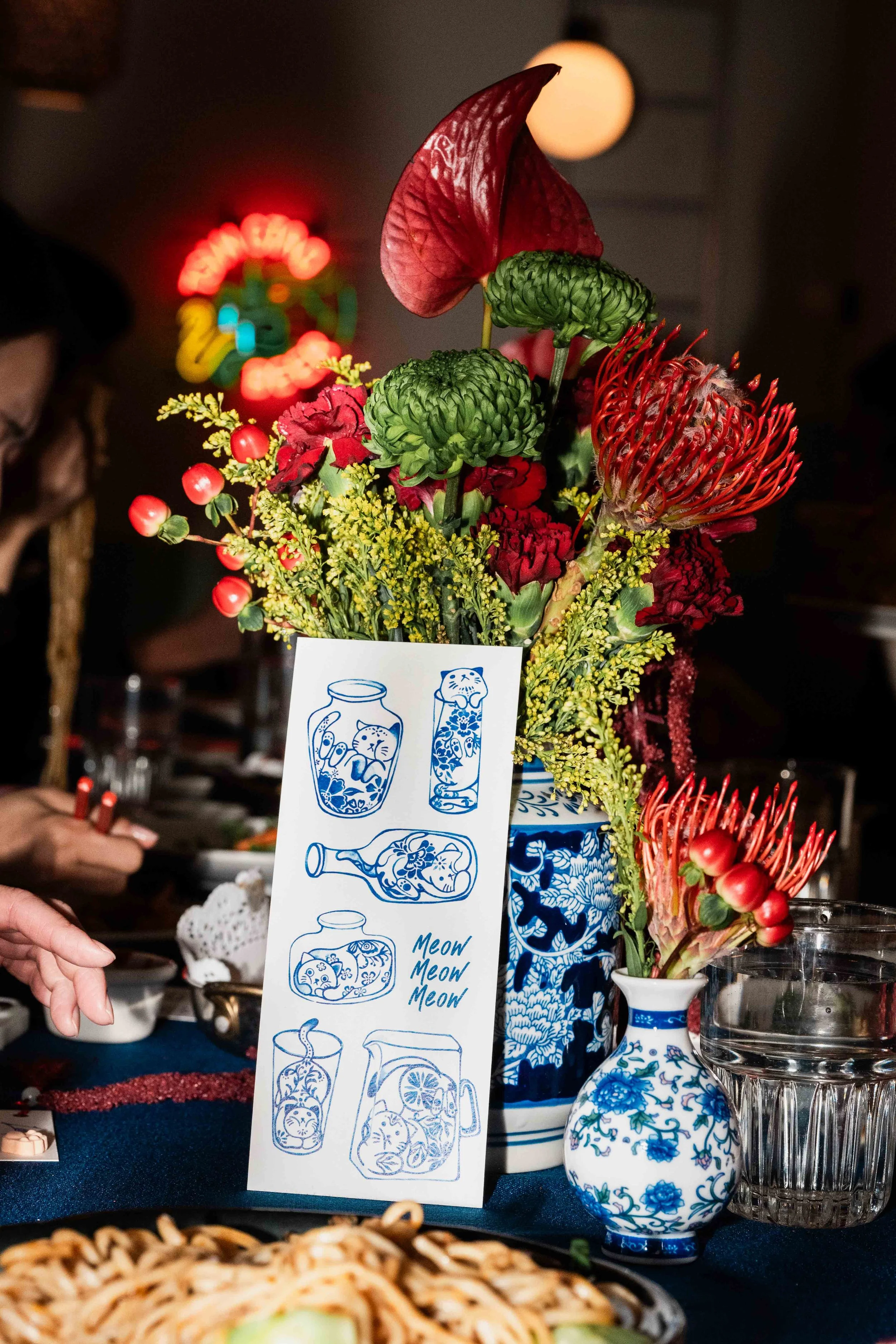 A floral arrangement with red, green, and yellow flowers in a blue and white vase, set on a table with a glass of water nearby. There is a sign with illustrations of various jars and the text "Meow Meow Meow." In the background, a lamp and some blurred neon lights are visible, indicating a dimly lit indoor setting.