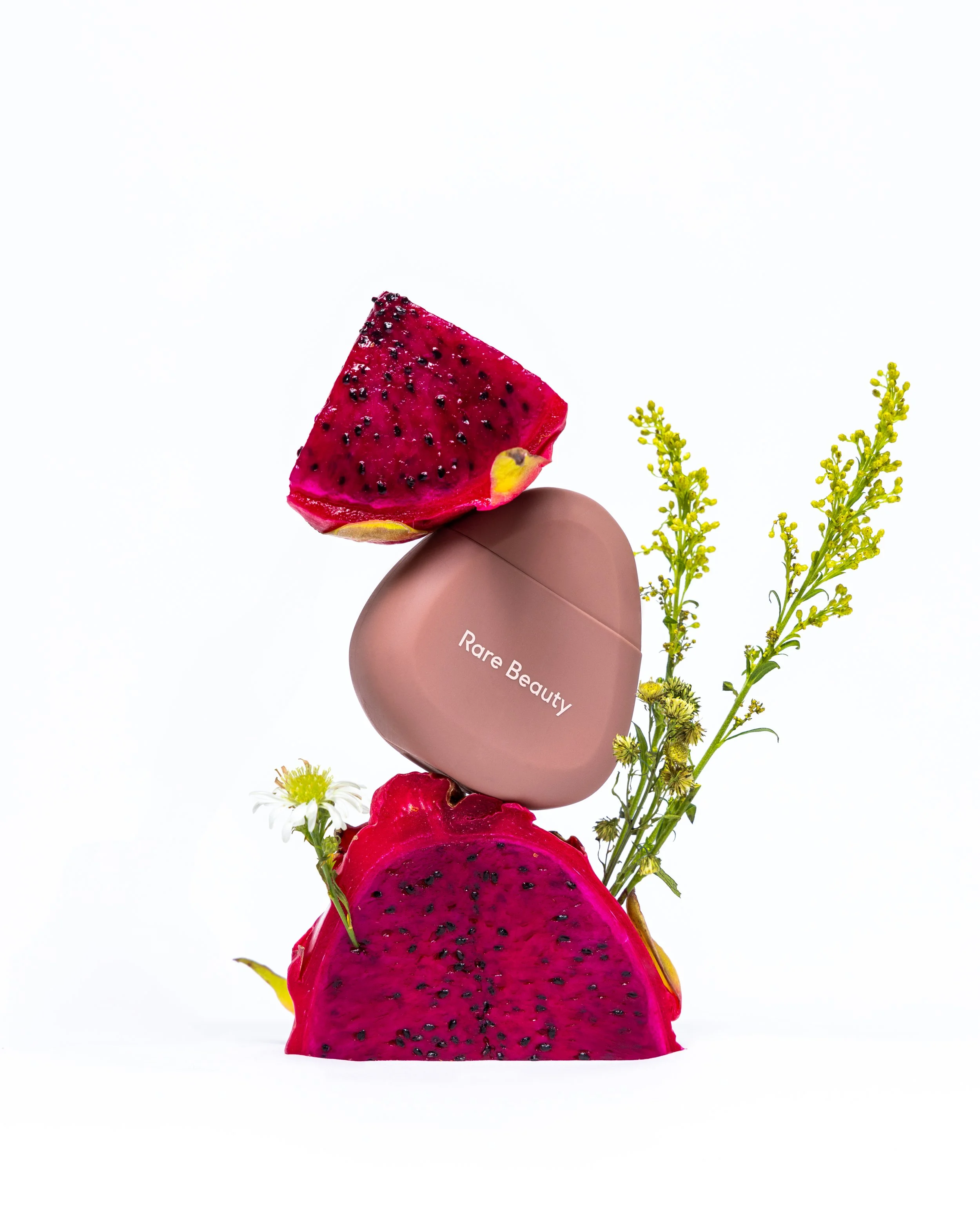 A small plant with yellow flowers and white blooms next to a pink dragon fruit sliced in half, with a beige oval object labeled "Rare Beauty" resting on top of it, against a plain white background.