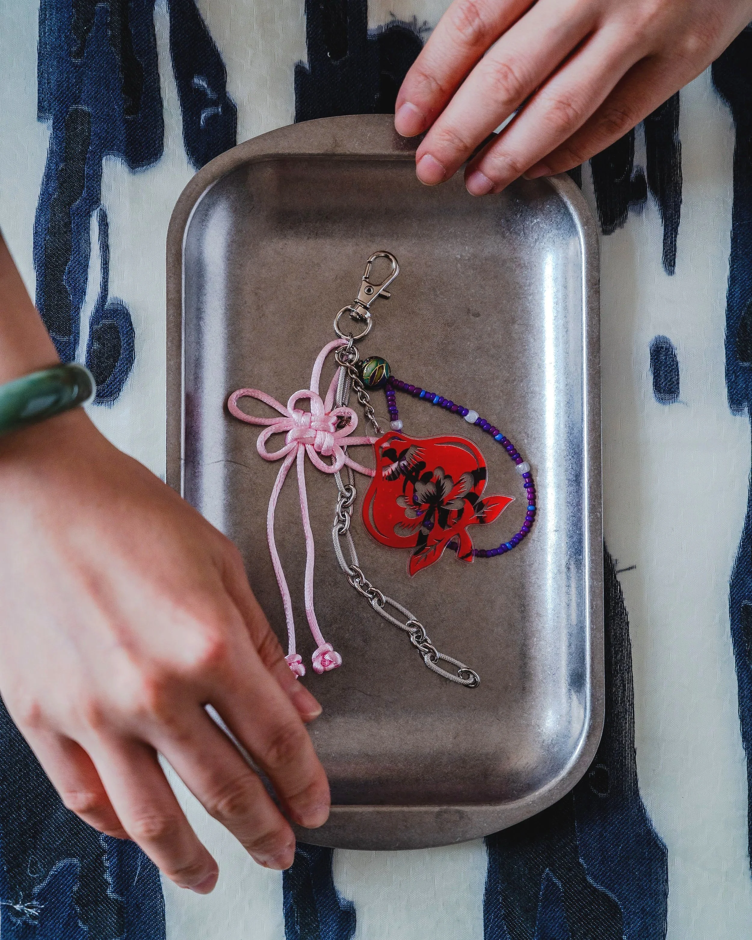 Close-up of a hand reaching into a metal tray containing various jewelry pieces, including a pink knotted bracelet, a red floral pendant, a chain, and colorful beaded necklaces, with a patterned cloth underneath.