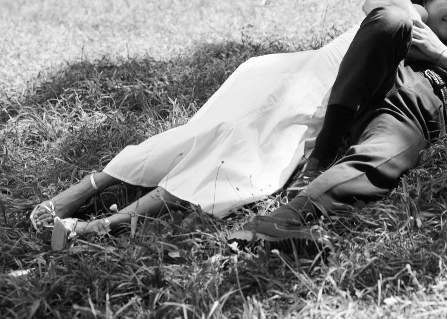 This is a black and white photo of a woman lying on the grass, dressed in a white dress and sandals, with a man beside her, partly visible, in a natural outdoor setting.