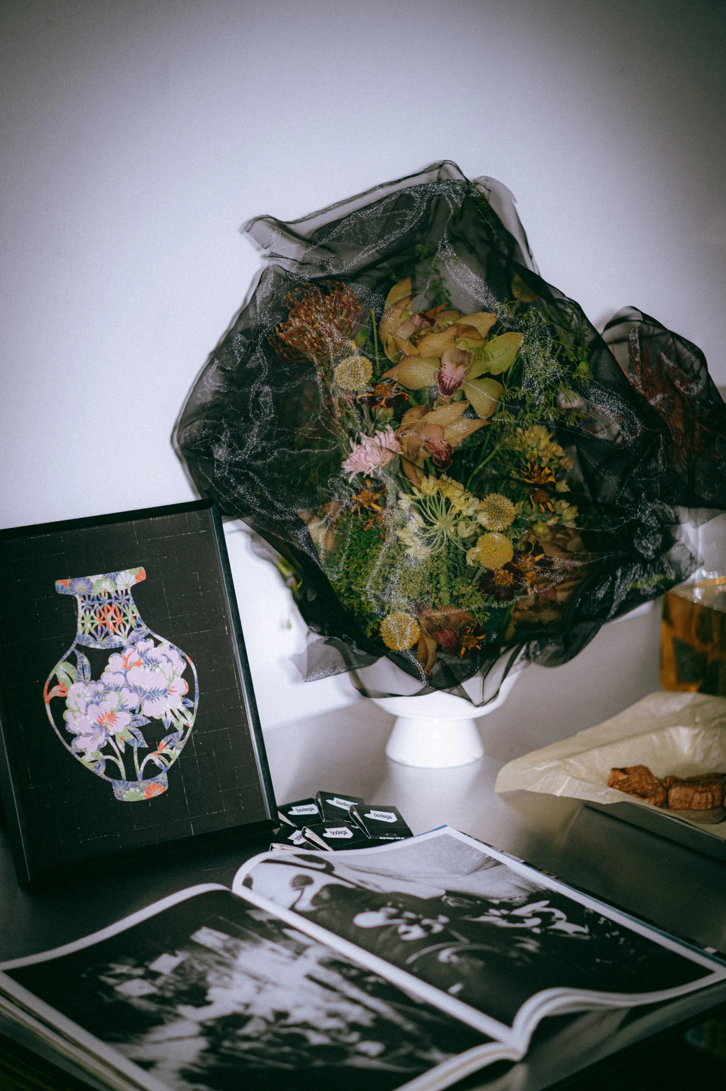 A bouquet of dried flowers wrapped in black transparent fabric is sitting on a white pedestal. Next to the bouquet, there is an open magazine displaying black and white photographs, and a black-themed image of a floral vase. There are also some small items with labels on the table.
