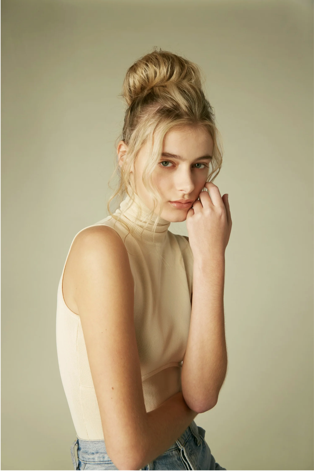 Young woman with blonde hair styled in a bun, wearing a sleeveless beige turtleneck top and light blue jeans, standing against a plain background, touching her face with her right hand.