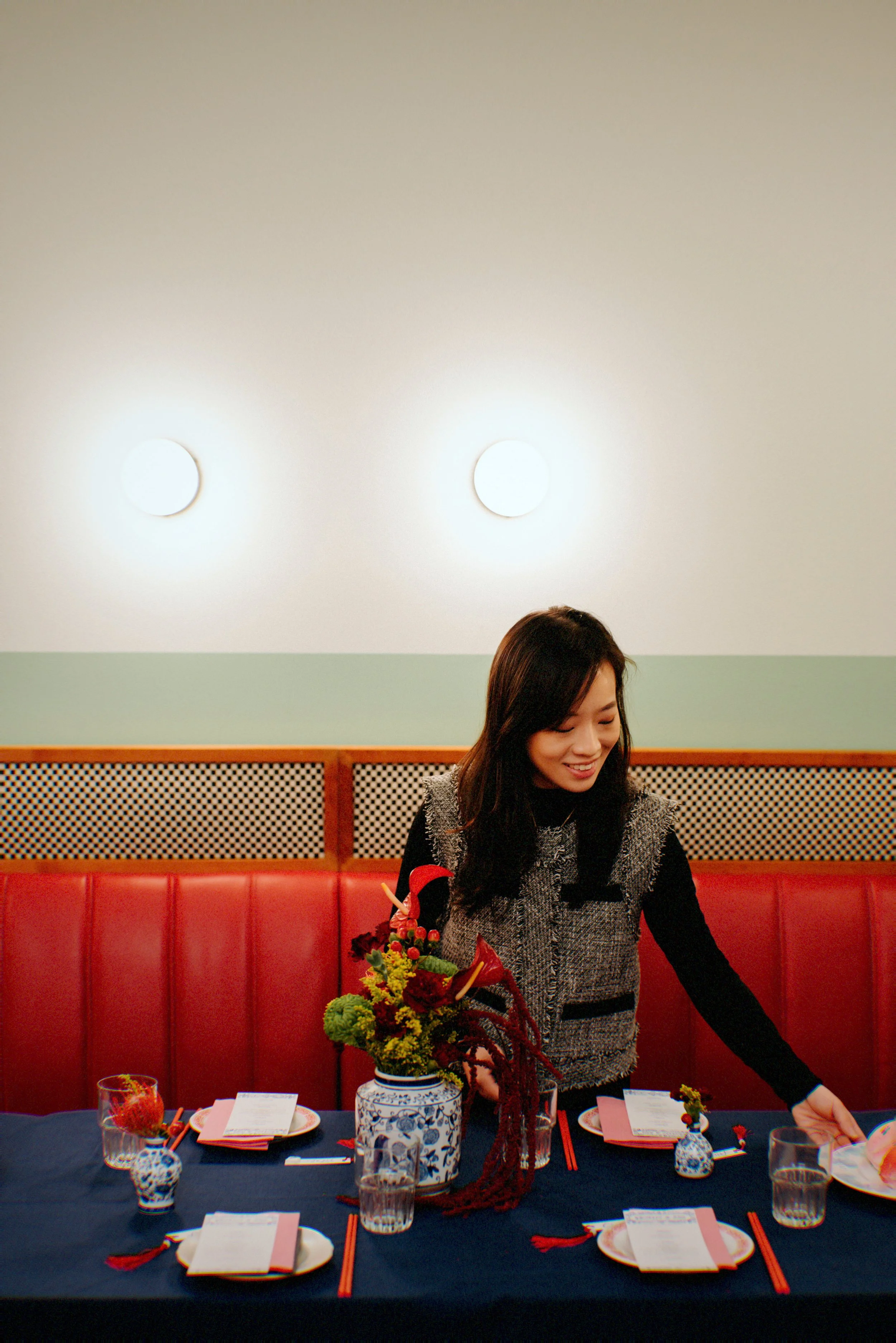 Woman setting a table with a centerpiece in a restaurant or dining room with a green and beige wall, two lights on the ceiling, and a red booth.