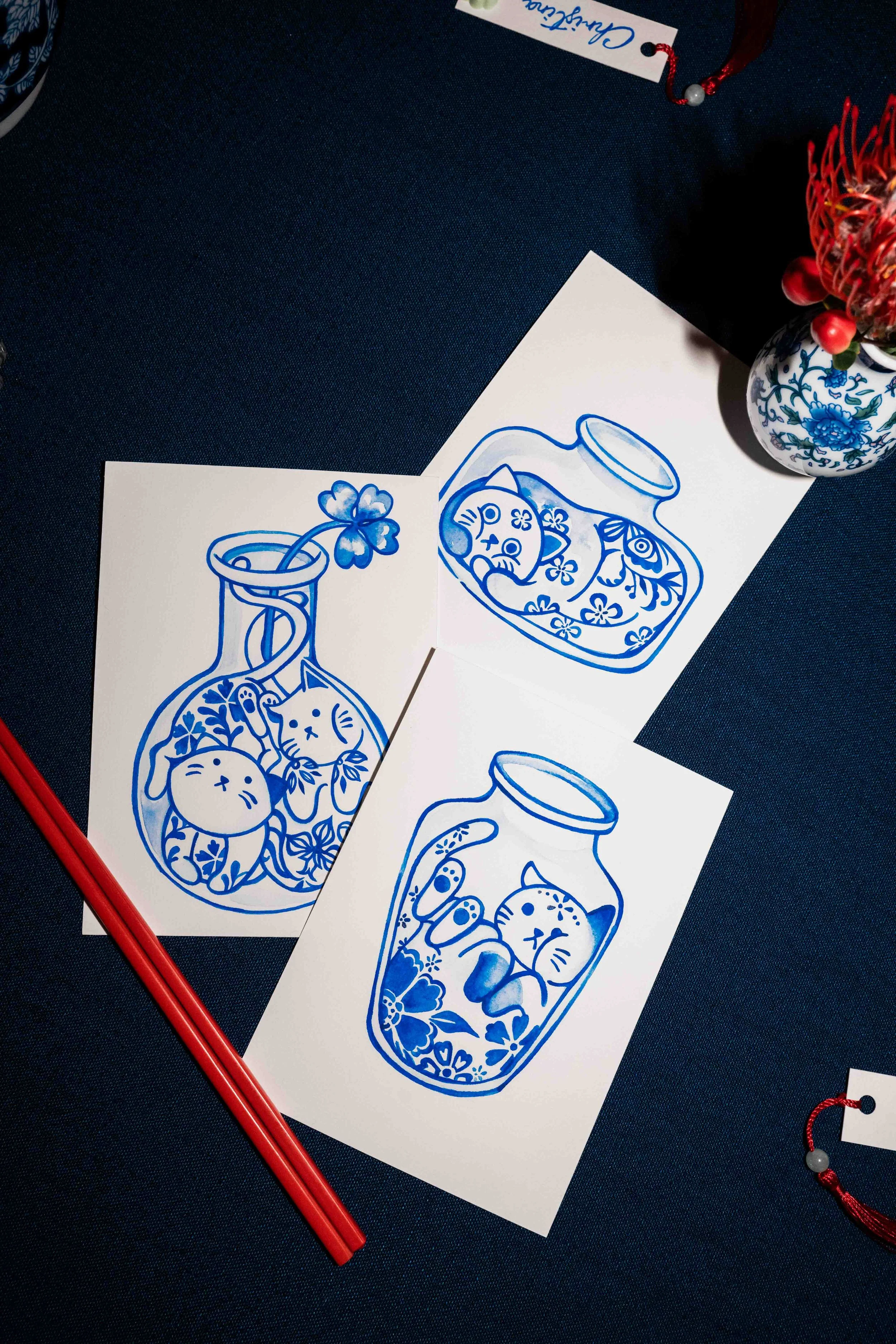 Three sheets of paper with blue illustrations of jars containing cats, flowers, and other decorative elements on a dark surface.