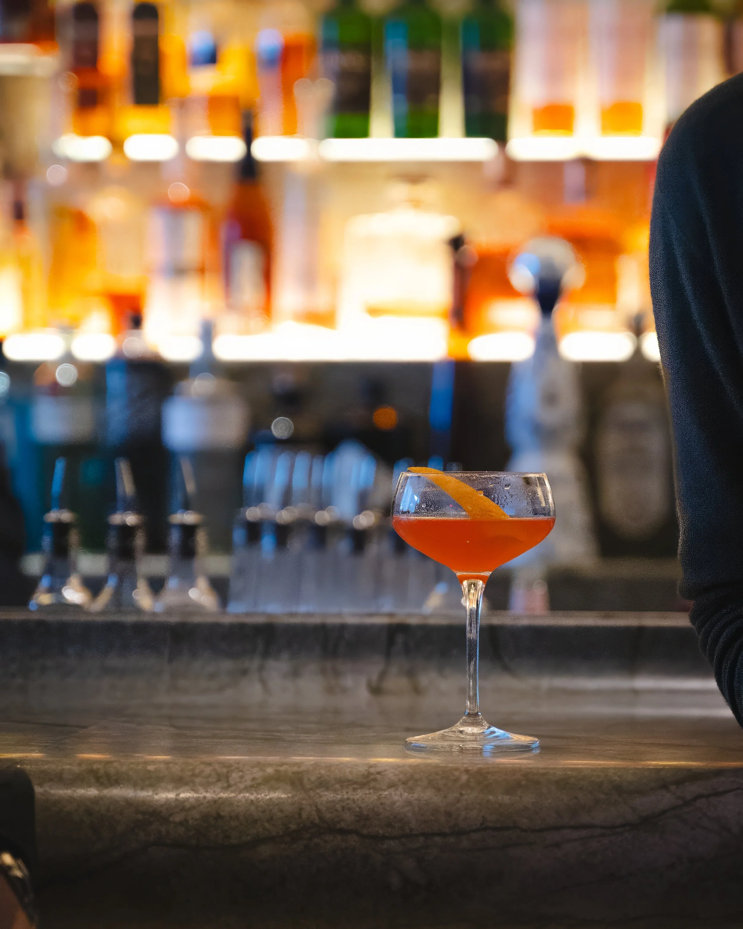 Close-up of an orange cocktail in a coupe glass with an orange twist garnish, placed on a bar counter with a blurred background of bottled drinks.