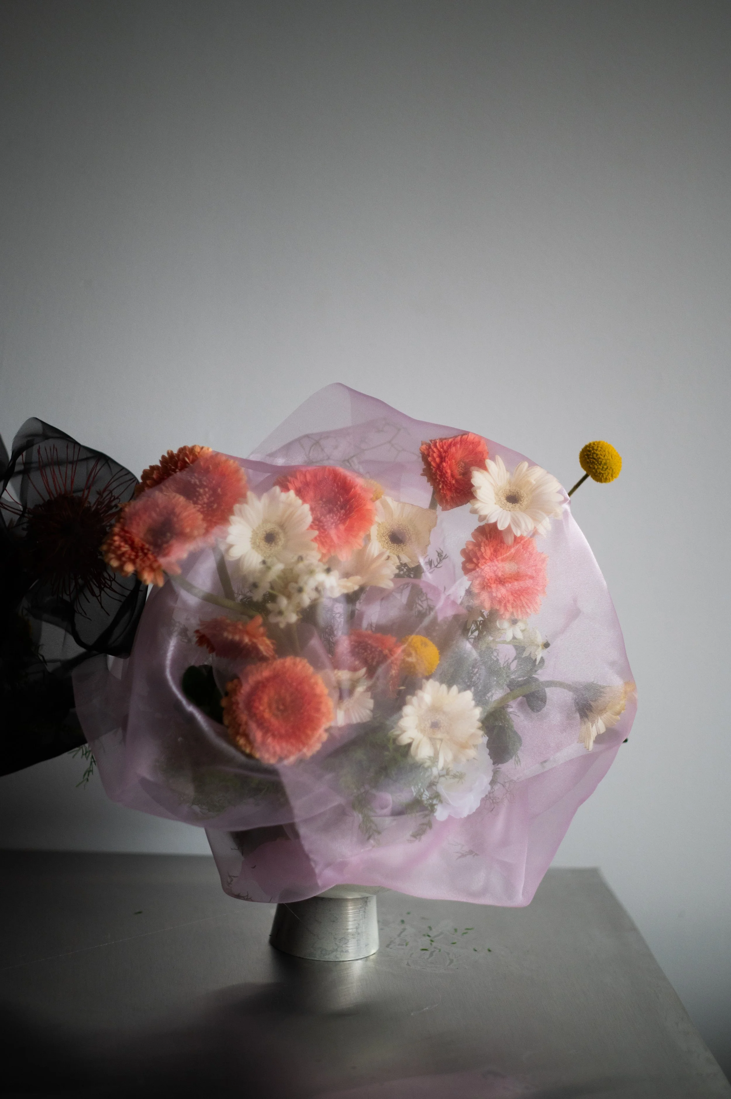 Colorful bouquet of flowers, including white daisies and orange and yellow flowers, wrapped in pink tissue paper, placed on a gray surface against a plain gray background.