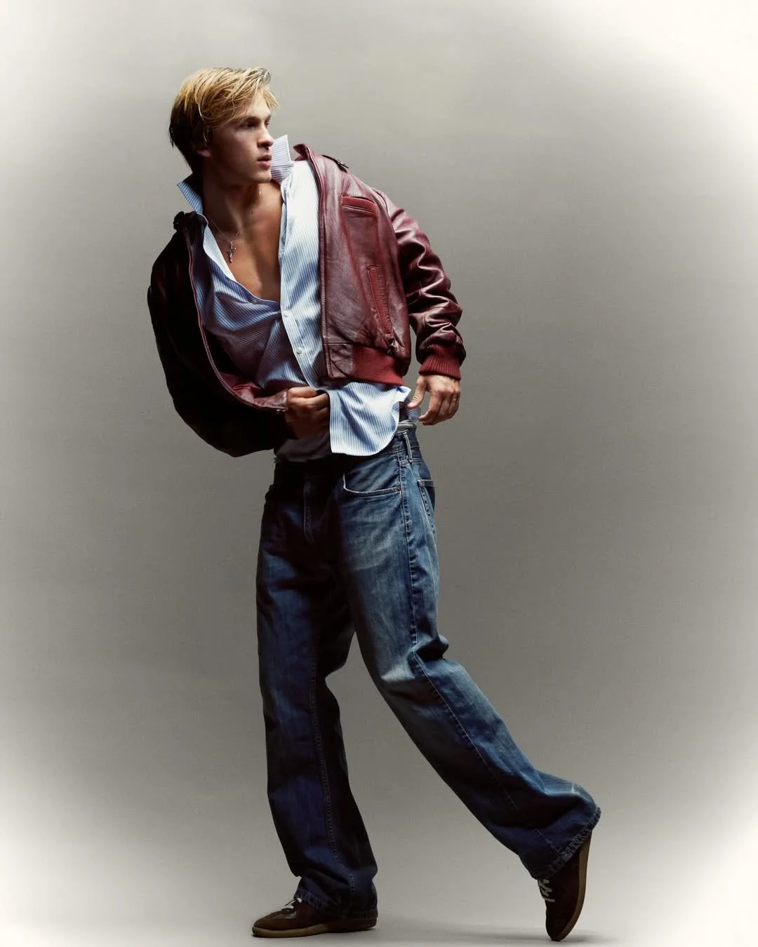 Young man with blond hair wearing a red leather jacket, unbuttoned blue and white striped shirt, and baggy blue jeans against a plain light gray background.