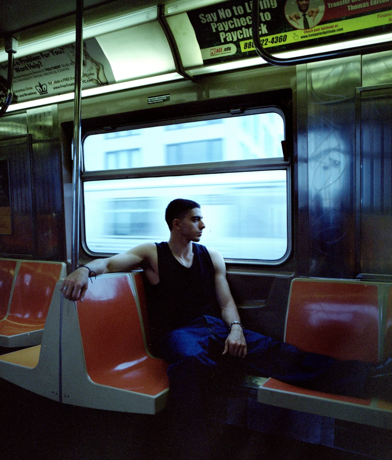 A young man sitting alone on an orange bench seat inside a moving subway train, looking out the window to his left. He is wearing a black sleeveless shirt and dark pants, with his right arm resting on the back of the seat and his left hand on his lap. The window shows a blurred cityscape passing by, indicating motion. Advertisements and signs are visible above the window.