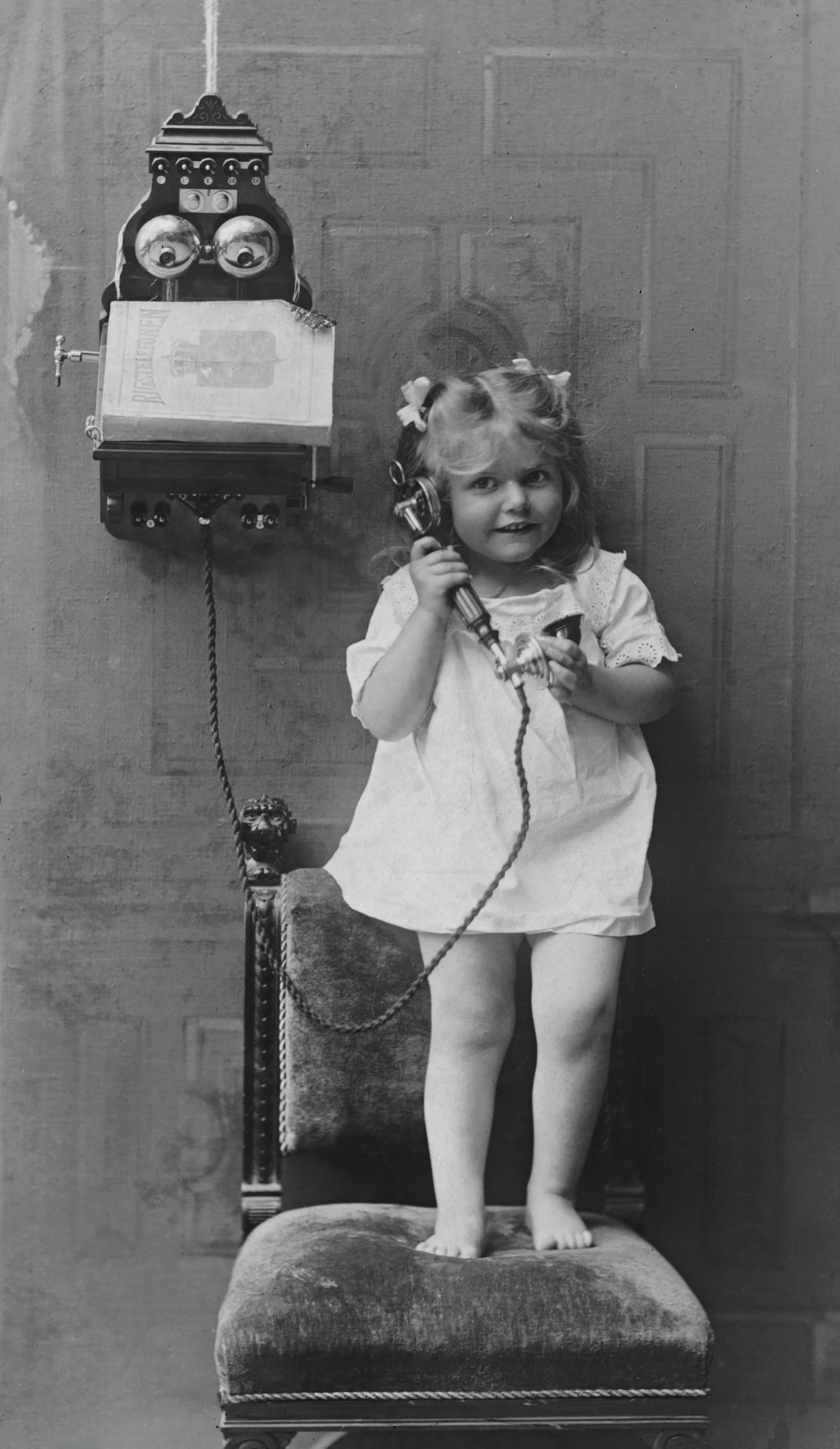 A young girl standing on a vintage chair, holding a rotary phone receiver to her ear, with an antique wall-mounted telephone with a box on top behind her.
