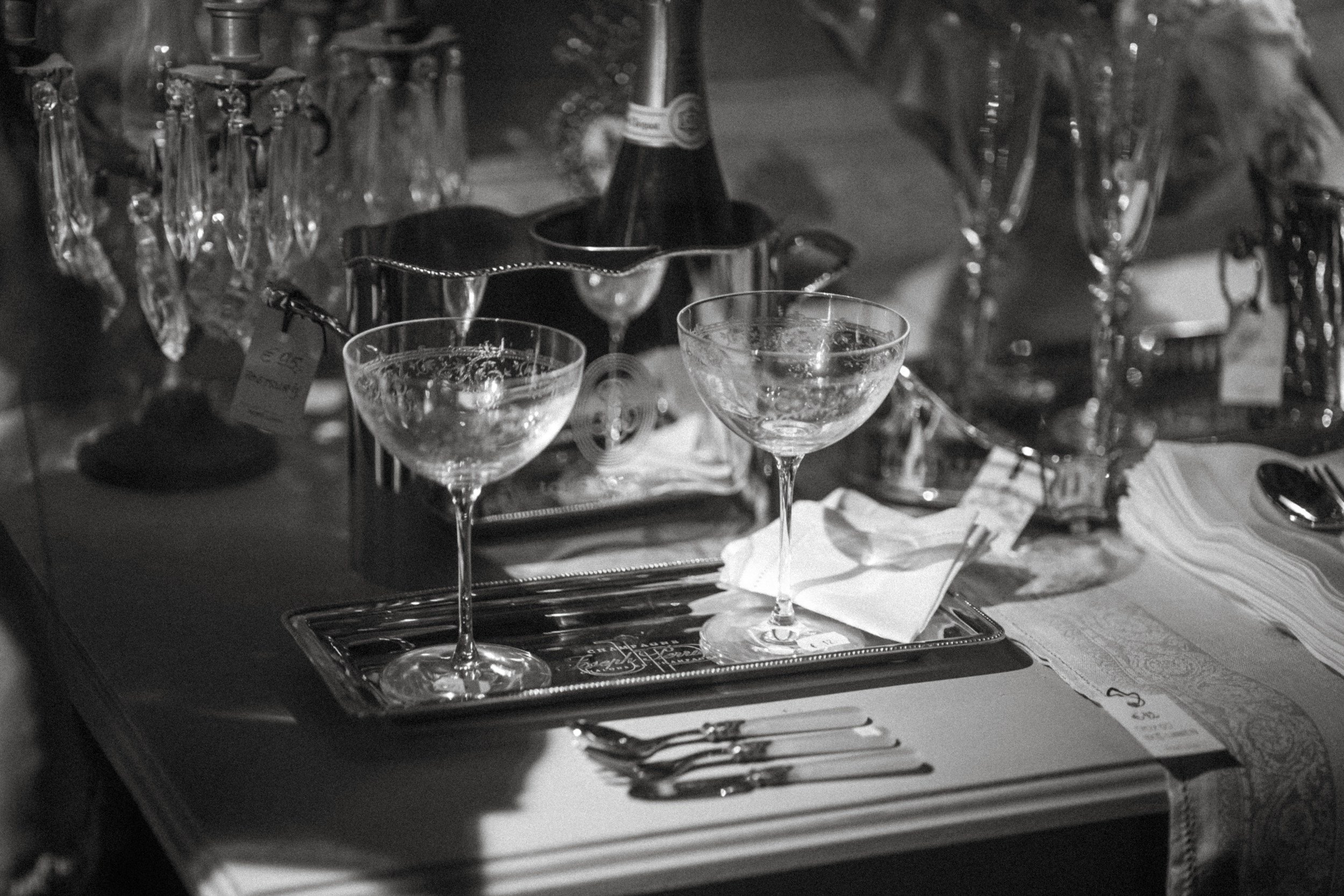A black and white photo of a table setting with two empty cocktail glasses, silverware, and various glassware items, with a tray and a bottle in the background.