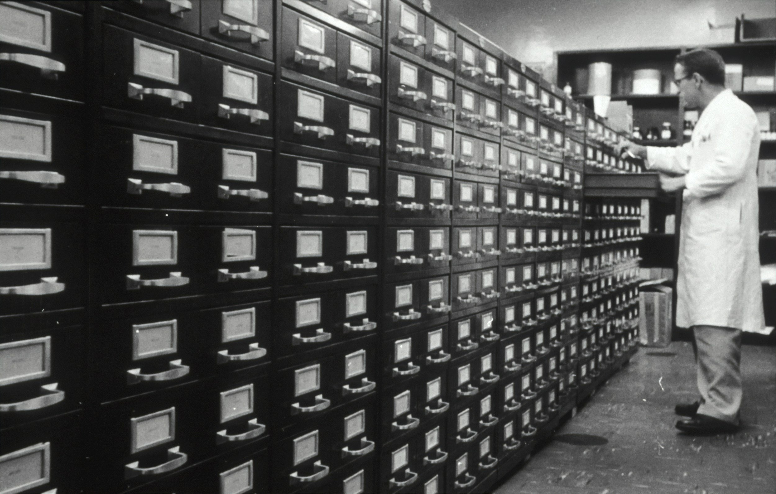 A man in a white coat working in a room with numerous filing cabinets.