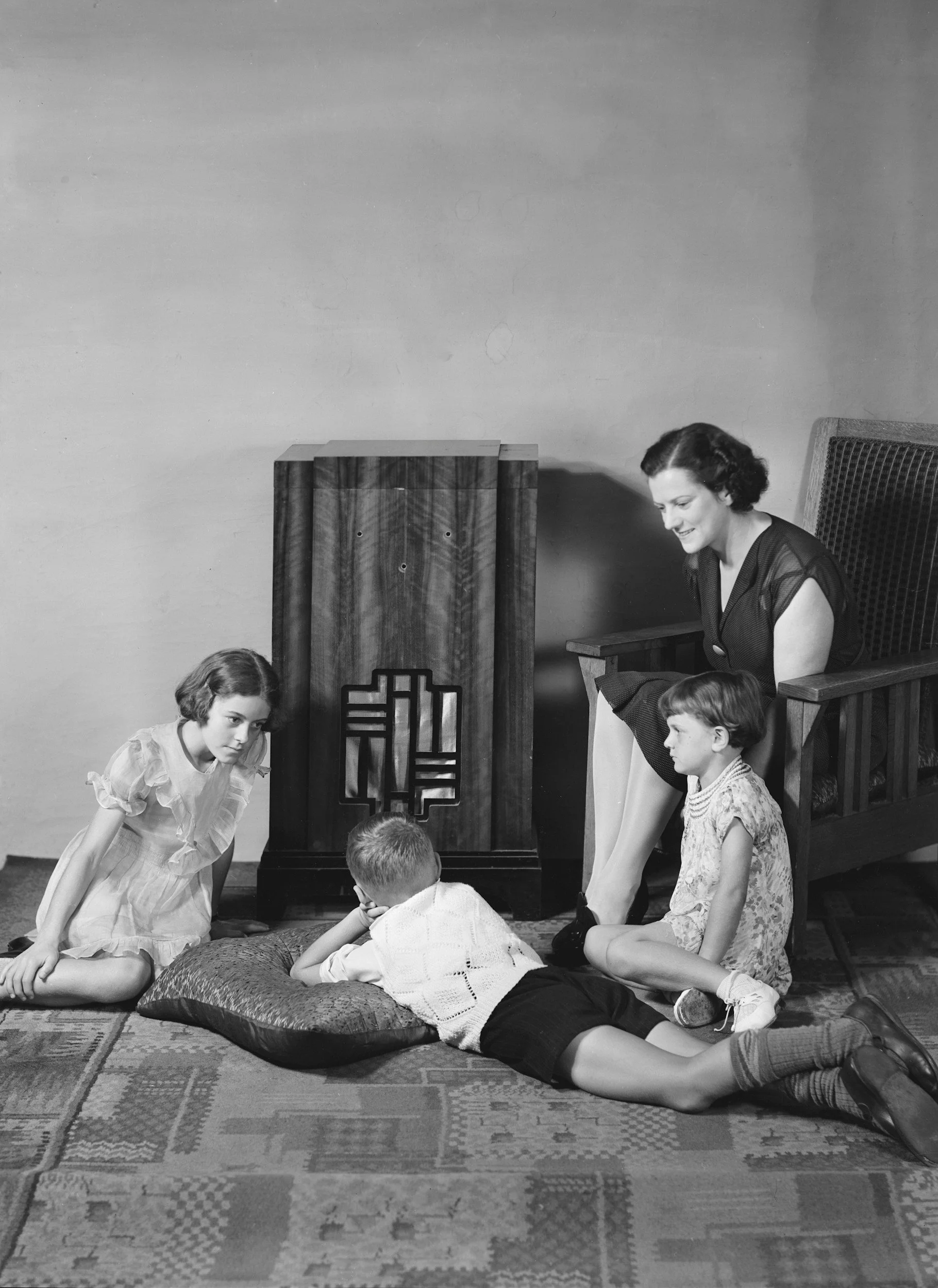 A woman and four children in a room, with the woman sitting on a large chair. The children are gathered around a pillow on the floor, appearing to be engaged in a serious or curious conversation, with some leaning forward and one lying on the pillow.