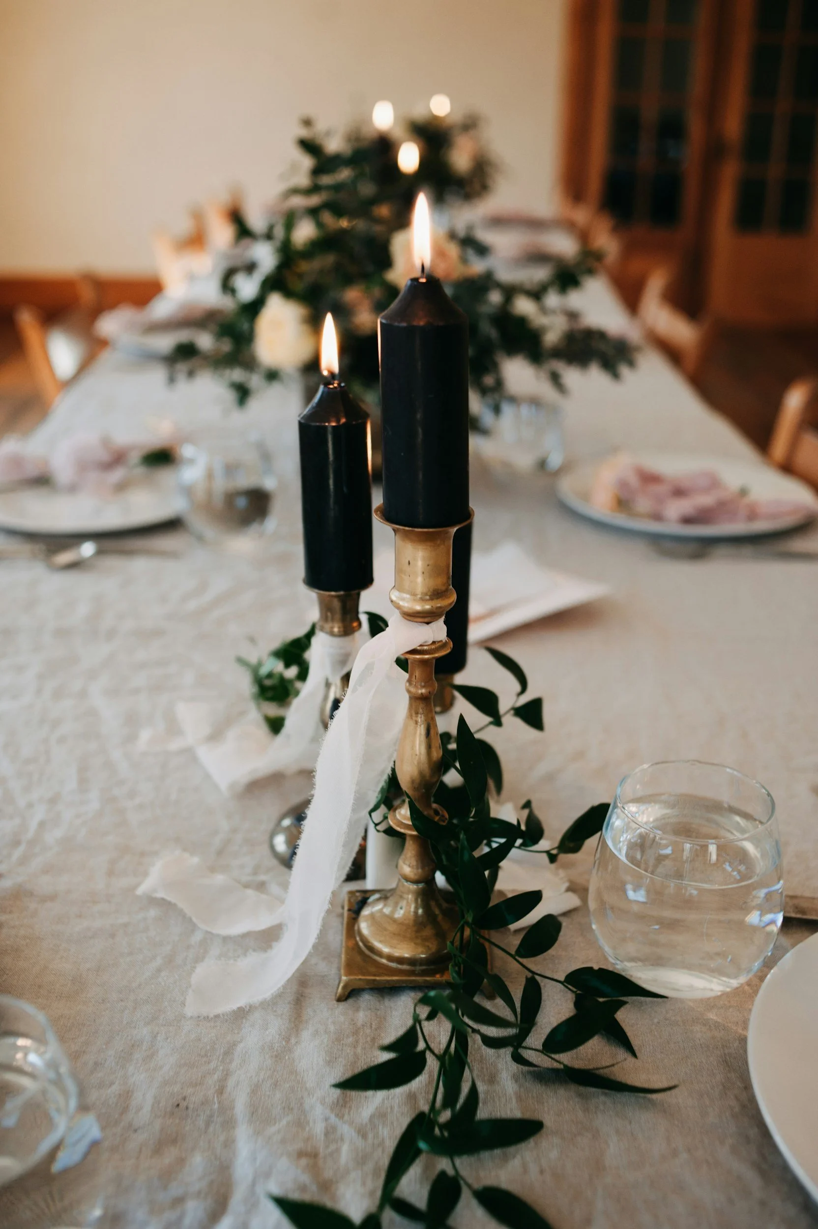 Elegant table centerpiece featuring three lit black candles in gold holders, decorated with green foliage, on a cream-colored tablecloth, with plates, glasses, and a blurred holiday-themed background.
