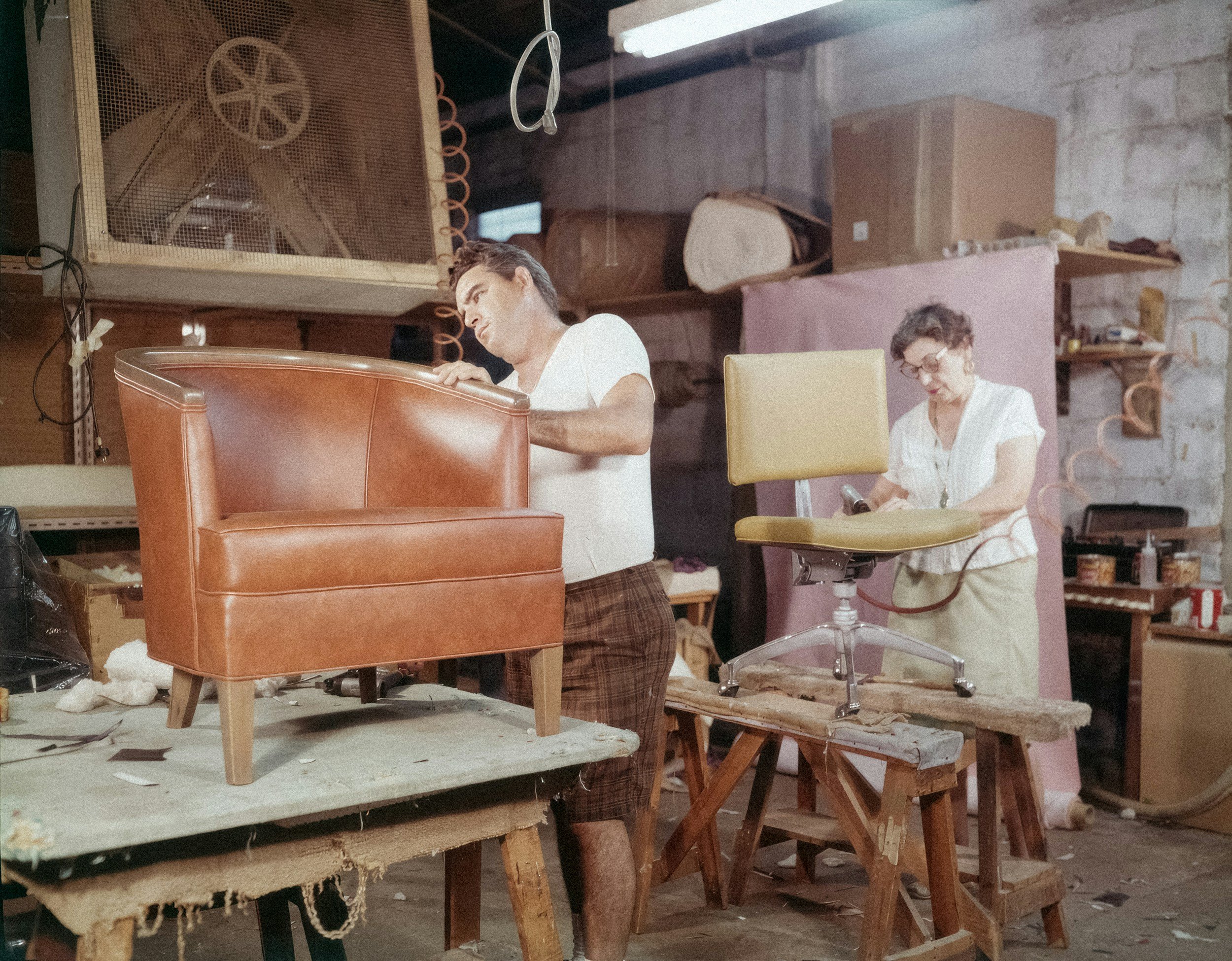 Two craftsmen working on furniture in a workshop; one is sanding a tan leather armchair, and the other is working on a yellow swivel chair.