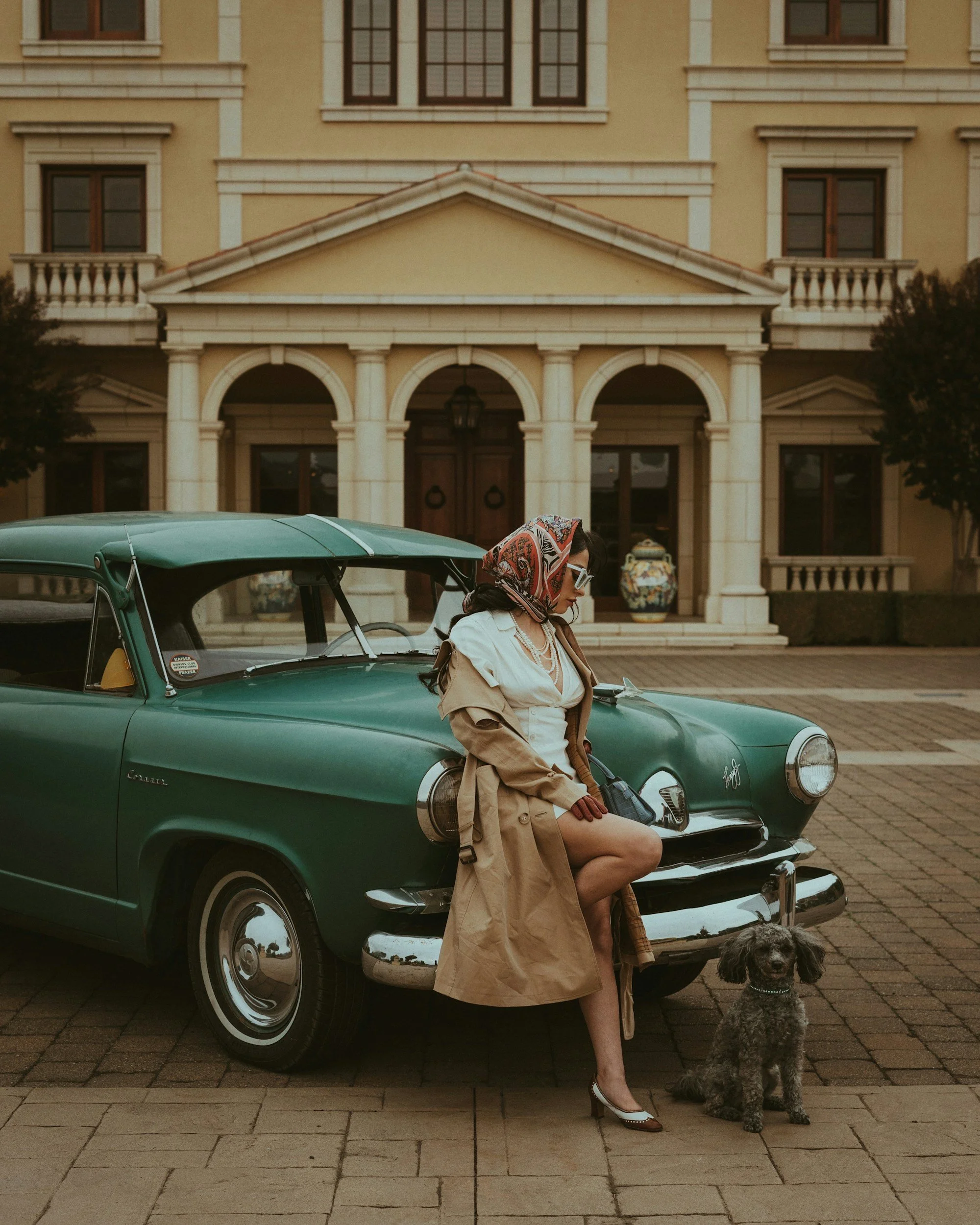 A woman sitting on the hood of a vintage green car parked in front of a grand yellow mansion, with a dog sitting on the ground beside her.