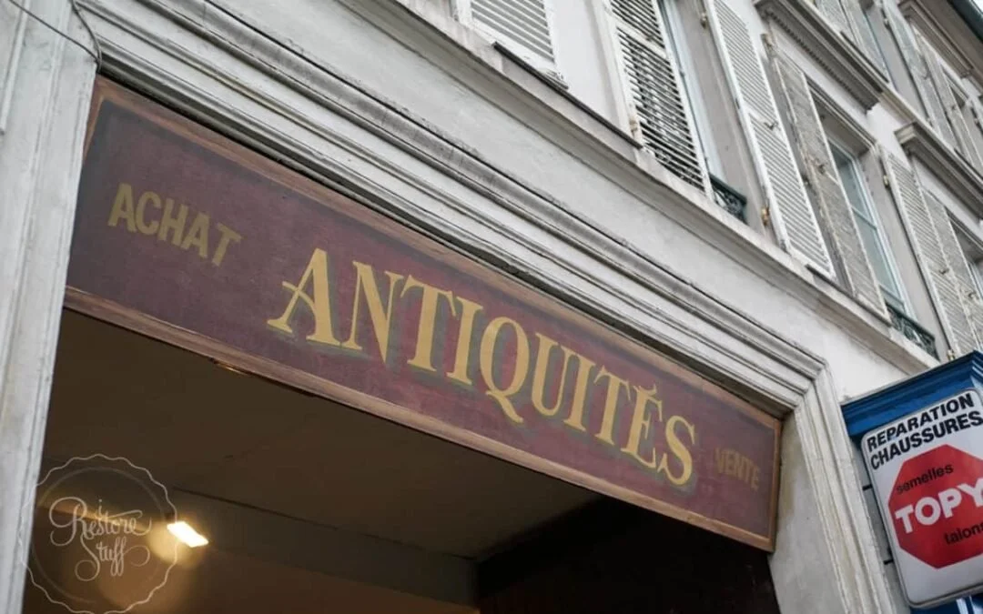 A vintage store sign reading "ACHAT ANTIQUITES" above the entrance of a building with French-style window shutters.