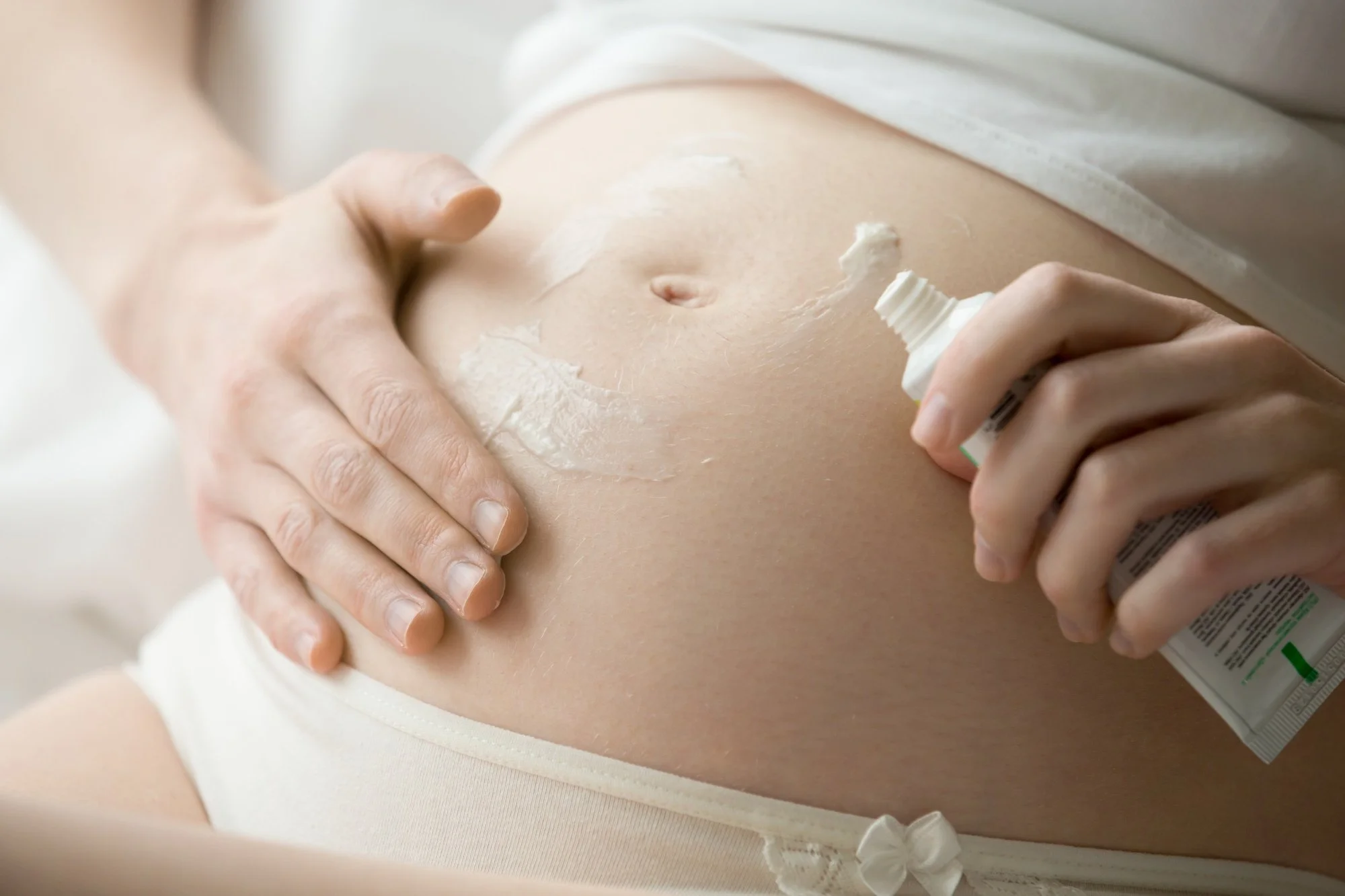 A pregnant person applying lotion or cream to their belly, wearing a white shirt and cream-colored underwear.
