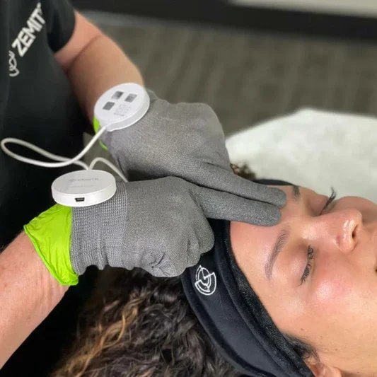 A woman receiving a facial treatment with a silver facial device applied to her cheek, lying down with a hair cap on at a spa or clinic.zemits neolifting