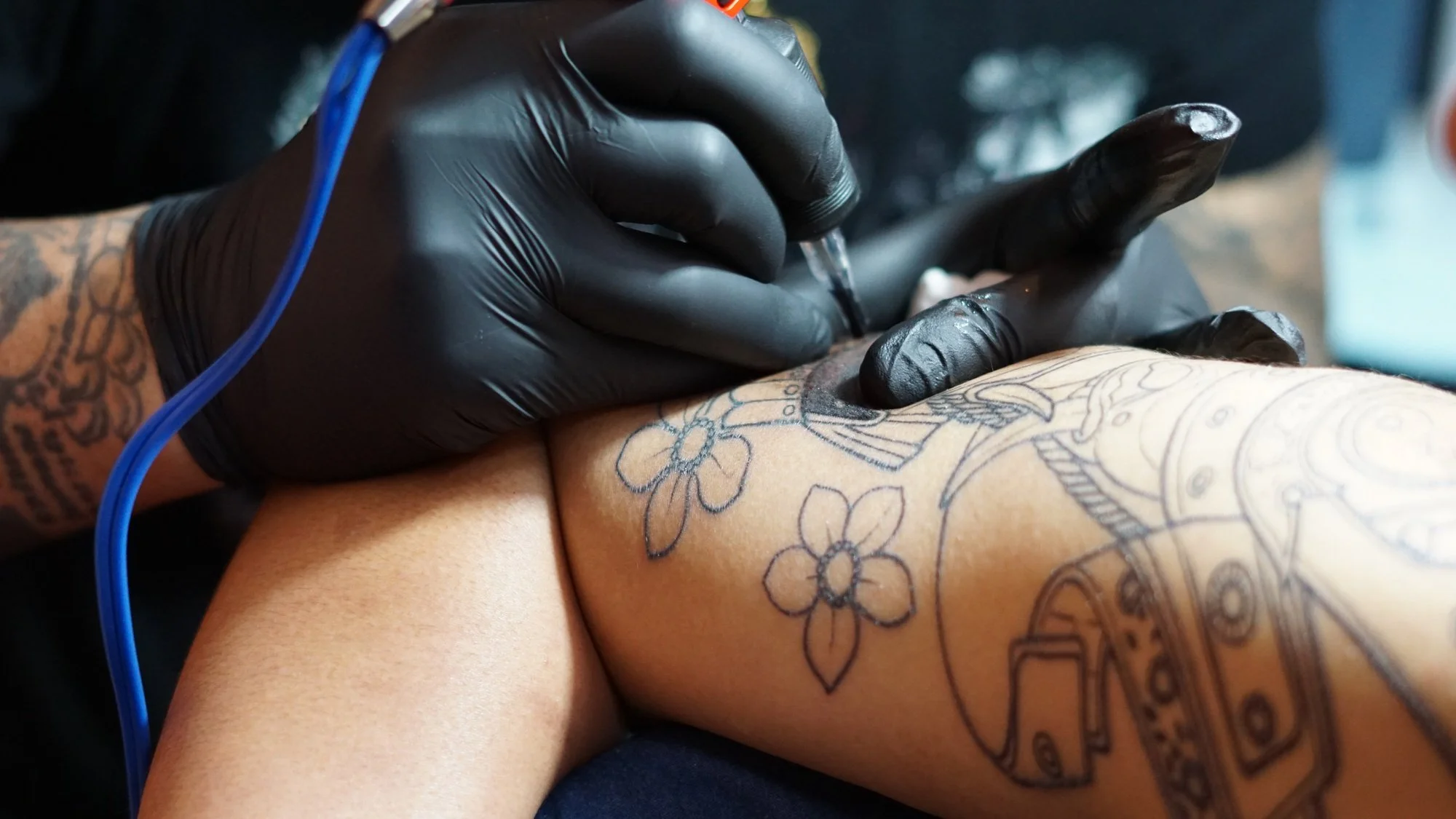 A tattoo artist wearing black gloves is tattooing a floral design on a person's thigh with a tattoo machine.