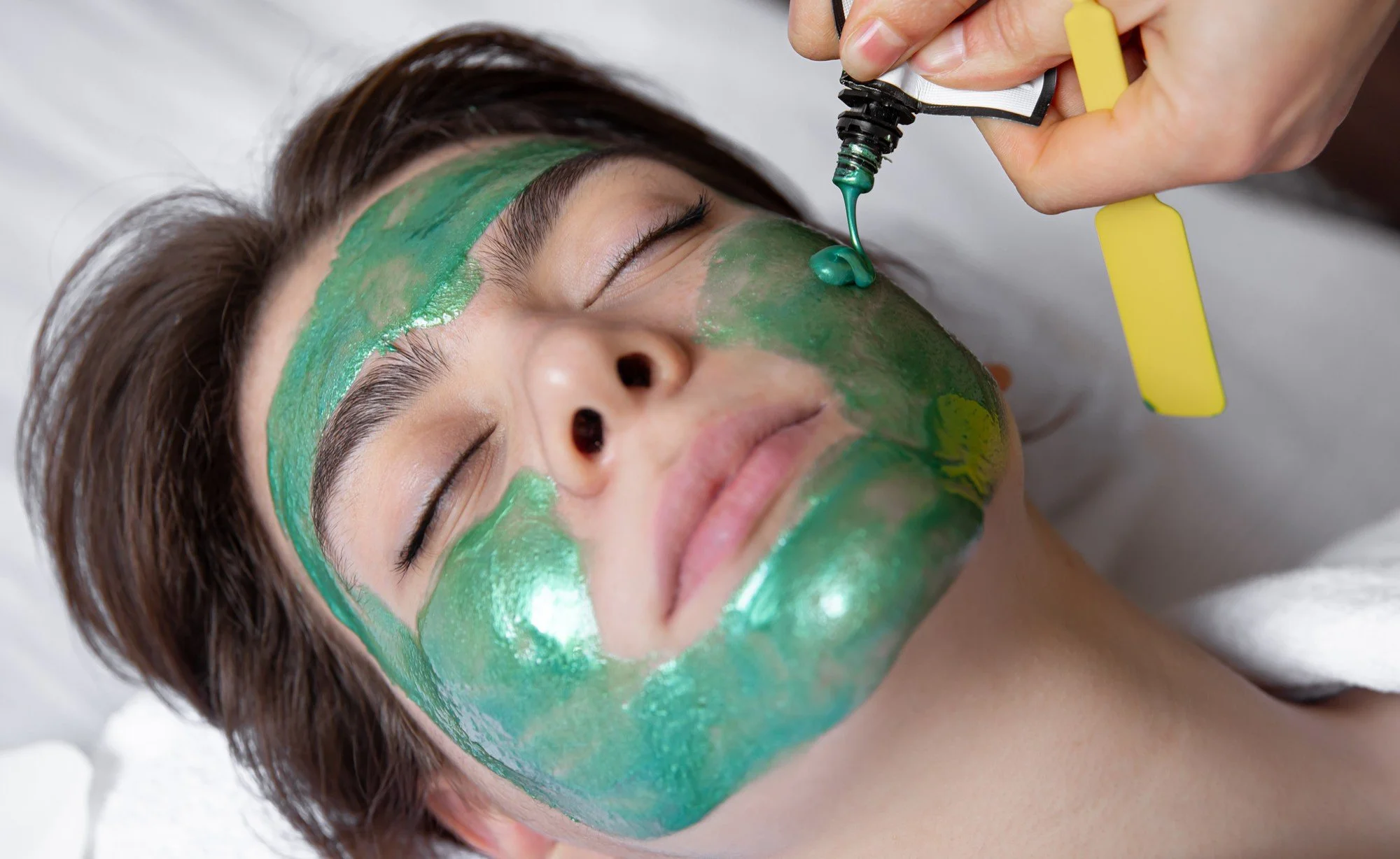 A woman receiving a green facial treatment while lying down with her eyes closed, and a skincare professional applying a green mask with a brush.