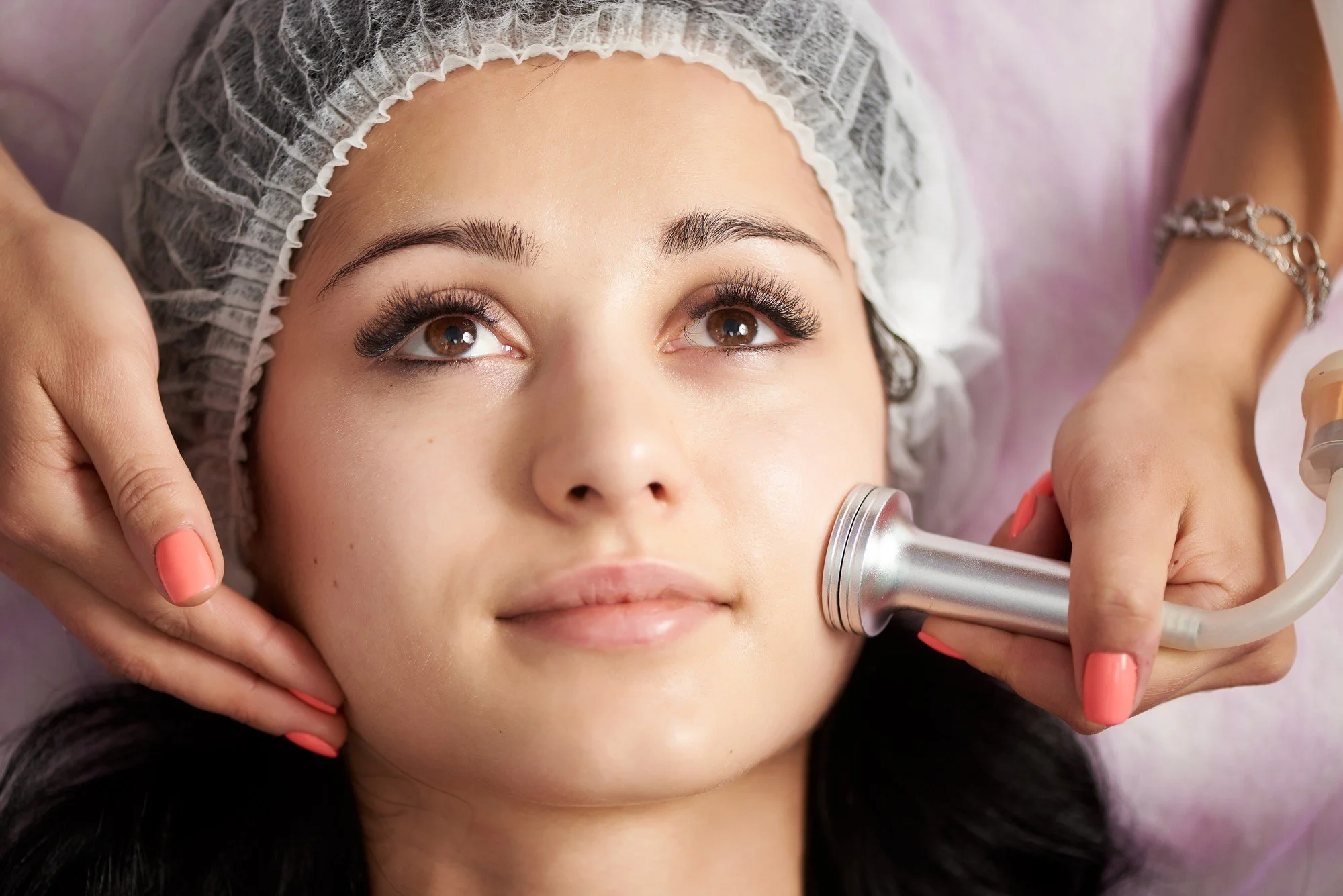 A woman receiving a facial treatment with a silver facial device applied to her cheek, lying down with a hair cap on at a spa or clinic.
