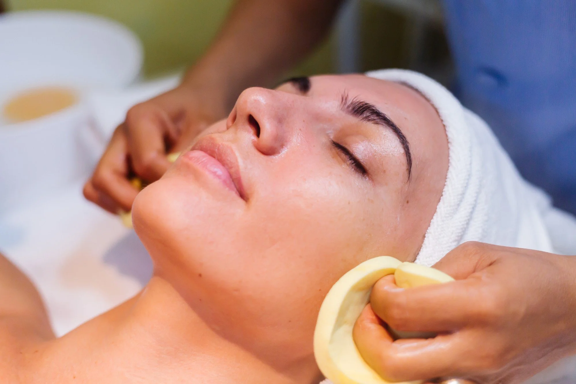 A woman with a white towel wrapped around her head receives a facial massage with a massage tool while lying down with her eyes closed.