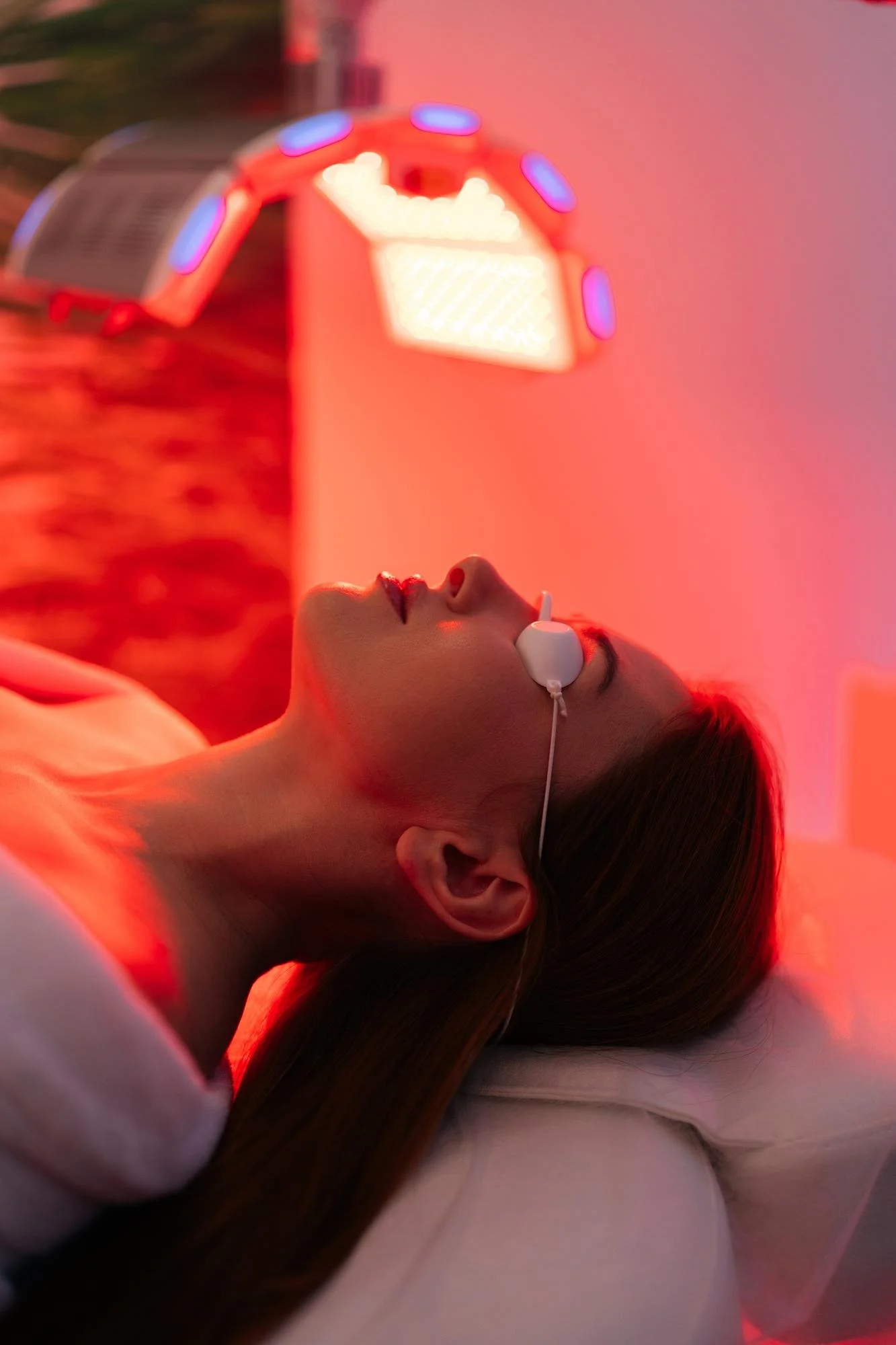 A woman lying down with her eyes closed, receiving red light therapy in a spa or wellness center, with a red and purple light in the background.