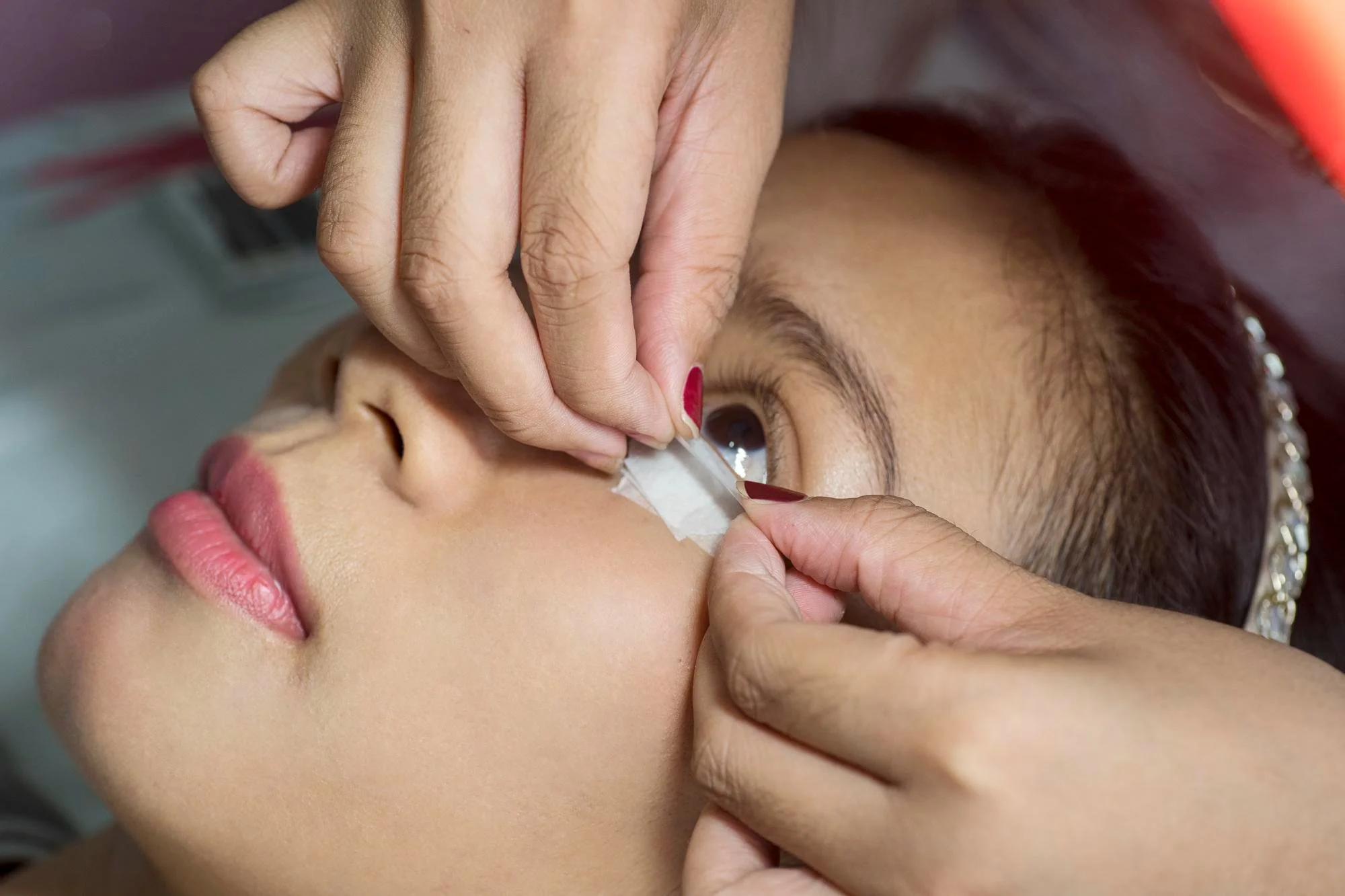 A woman with dark hair and fair skin getting a beauty treatment on her eye, with a technician applying a patch or tape under her eye.