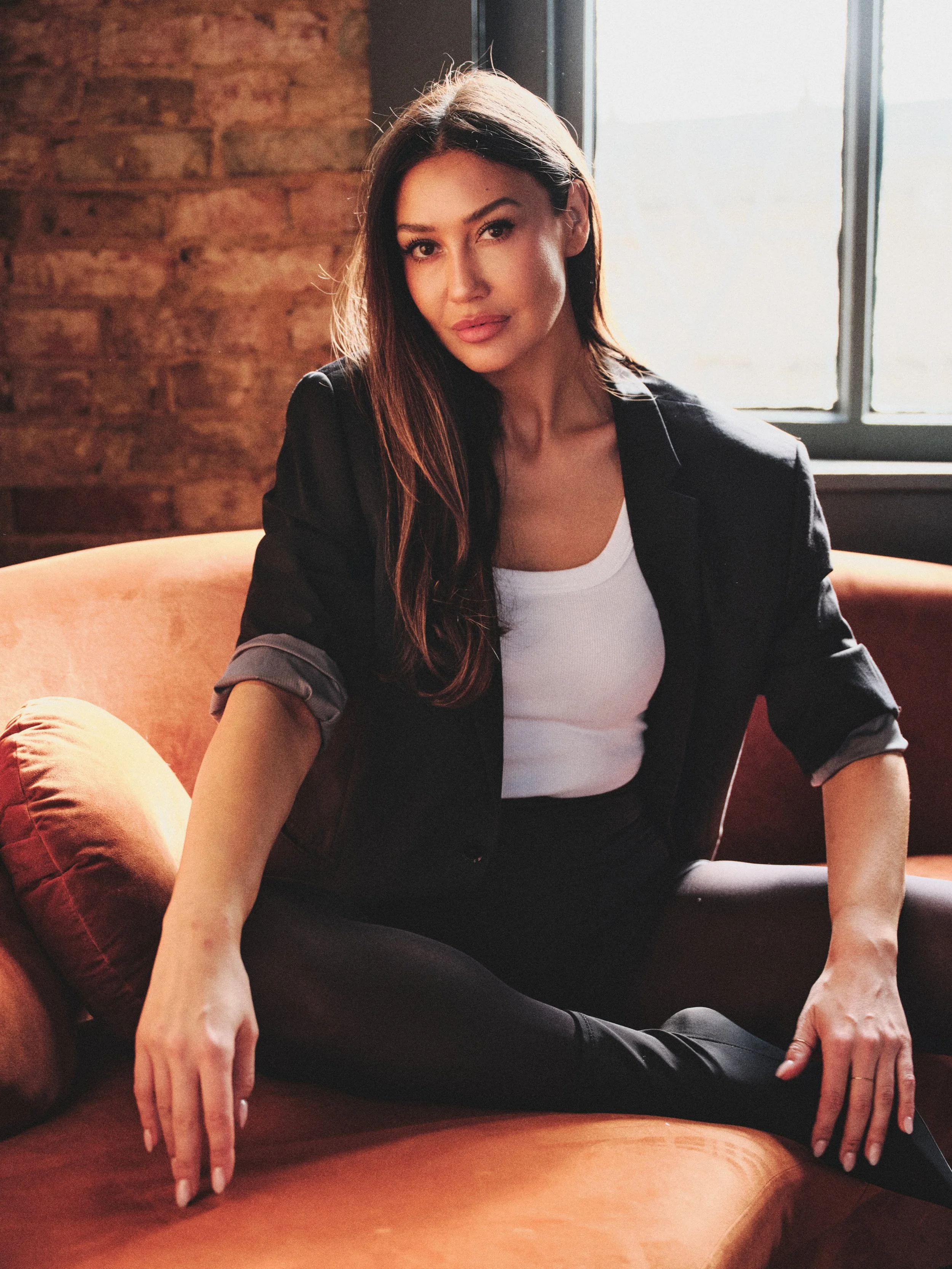 A woman sitting on a brown sofa near a window, with her legs crossed and one hand resting on the sofa. She has long brown hair, light makeup, and is wearing a black blazer over a white top, with black pants.