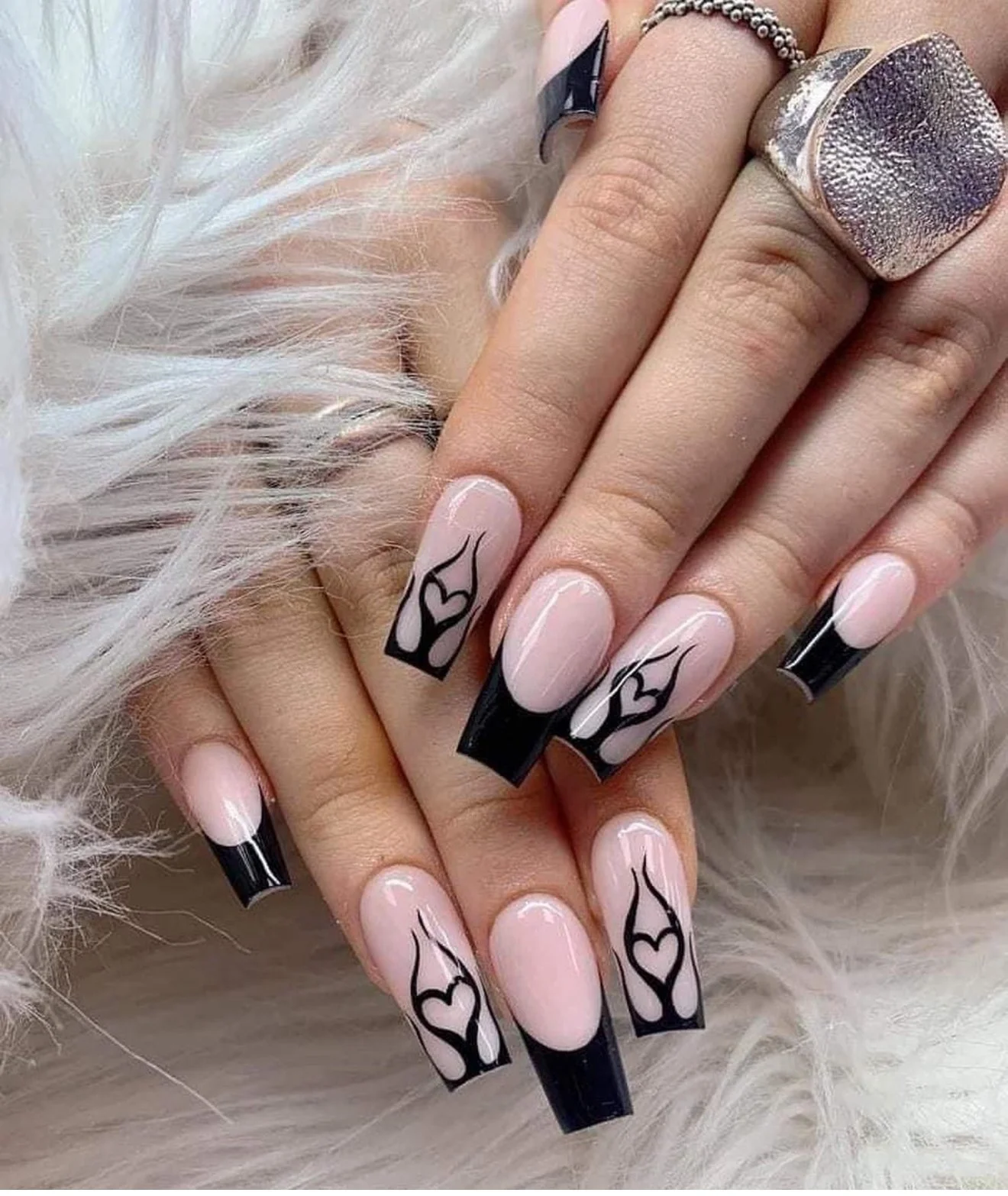 Close-up of hands with long acrylic nails. The nails are French manicured with black tips and black outline heart designs on a nude base. The person is wearing rings, including a large textured ring and a beaded ring, and has platinum blonde hair in
