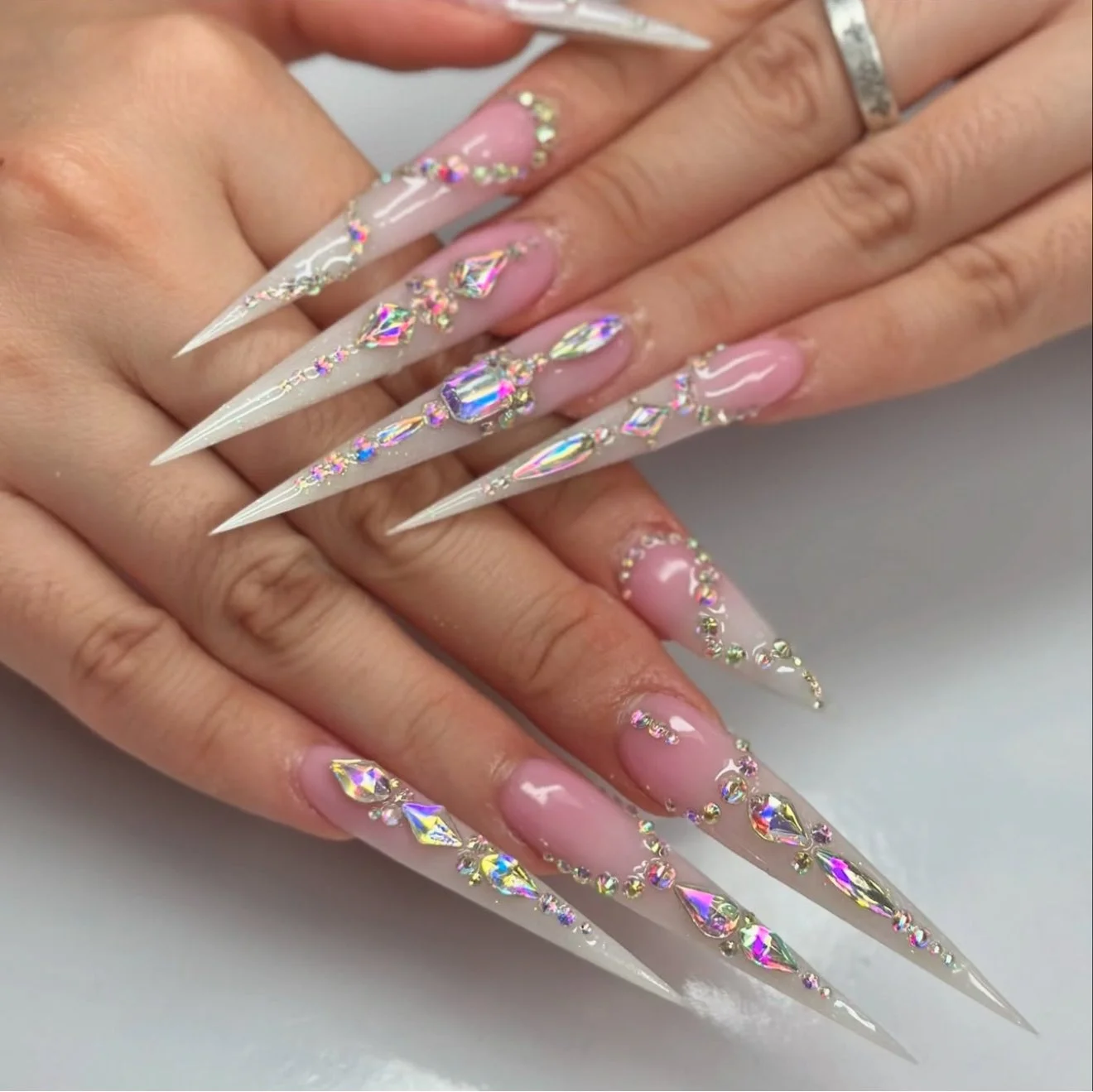 Close-up of long, pointed acrylic nails decorated with iridescent rhinestones and crystals, with a nude pink base polish, held by a person's hands.