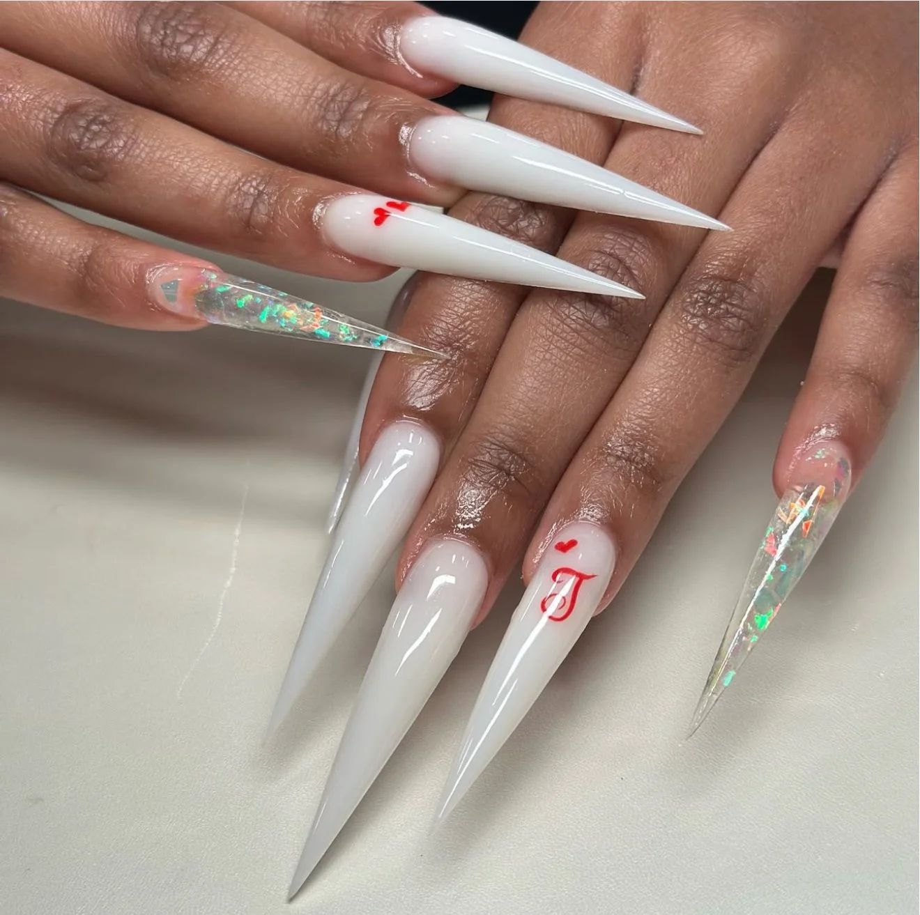Close-up of long, pointed white acrylic nails with red heart and letter designs, clear holographic accent nails, and manicured fingers resting on a white surface.