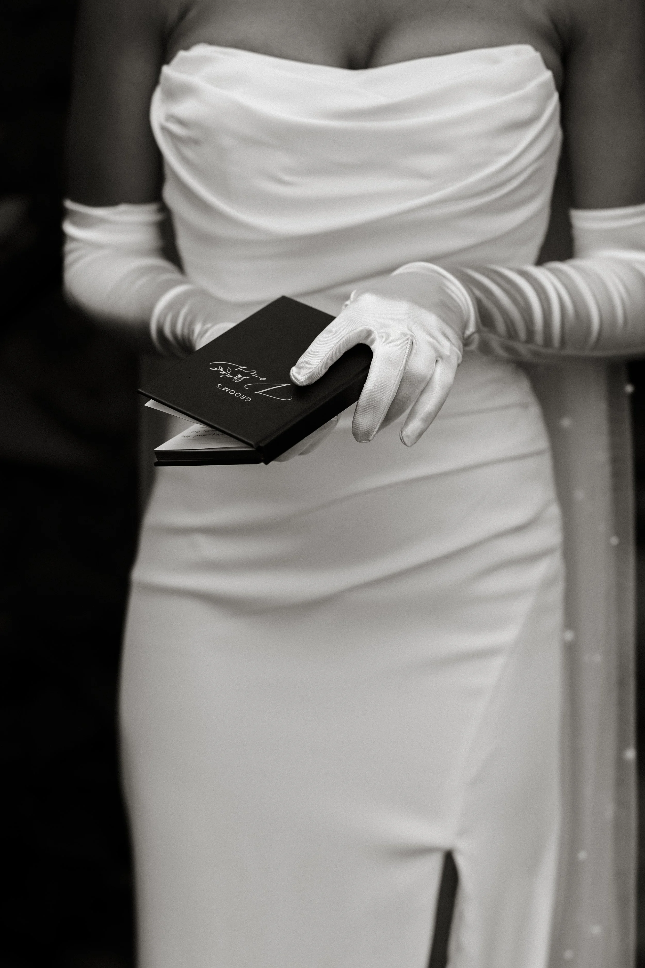 A woman dressed in a white wedding gown and gloves holding a black wedding program or booklet.