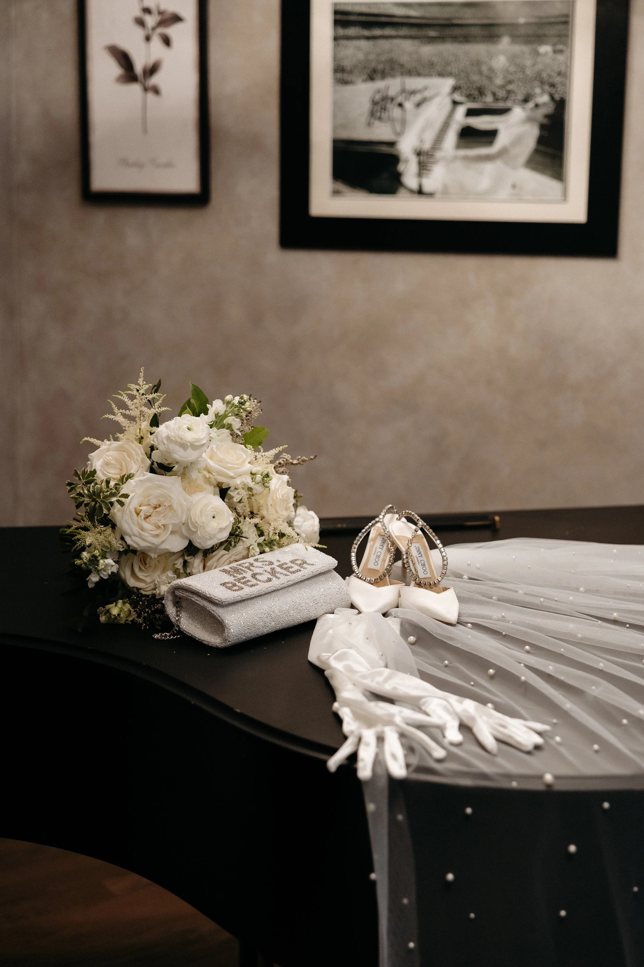 A wedding dress, bouquet of white roses, white gloves, and jewelry displayed on a black table.