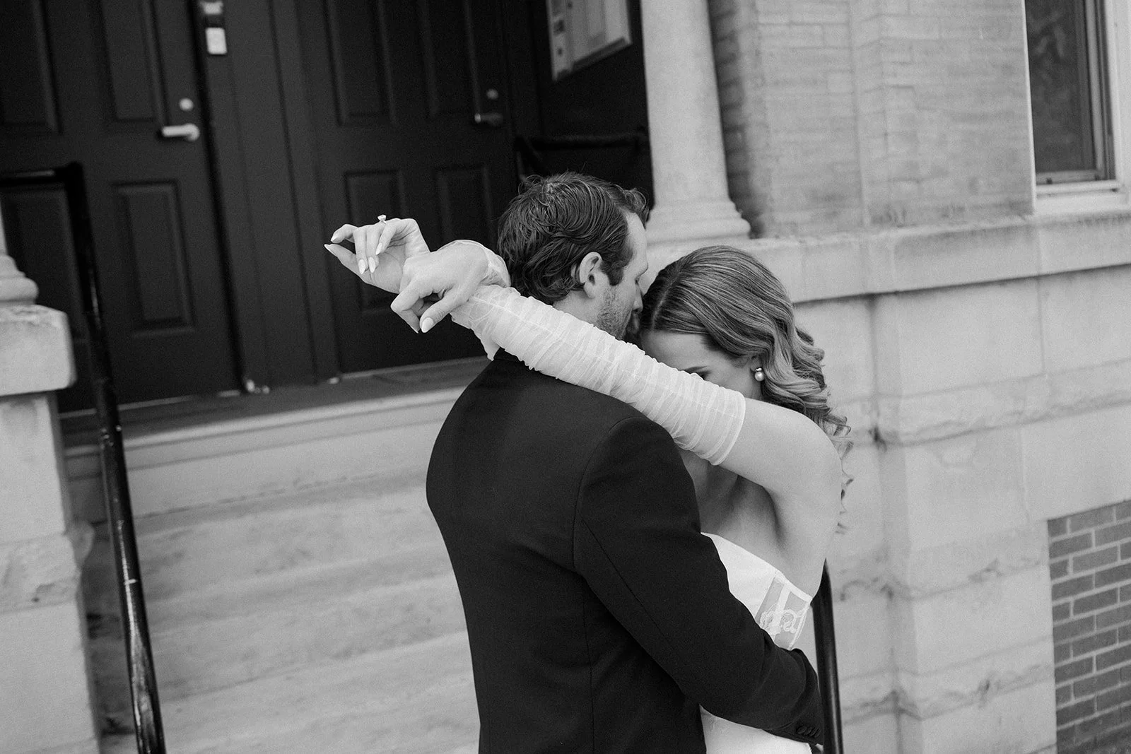 A black and white photo of a couple hugging outside, with the woman’s arms wrapped around the man and their heads close together. The woman is wearing a dress with long sleeves and earrings, and the man is in a suit. They appear to be in a tender, in