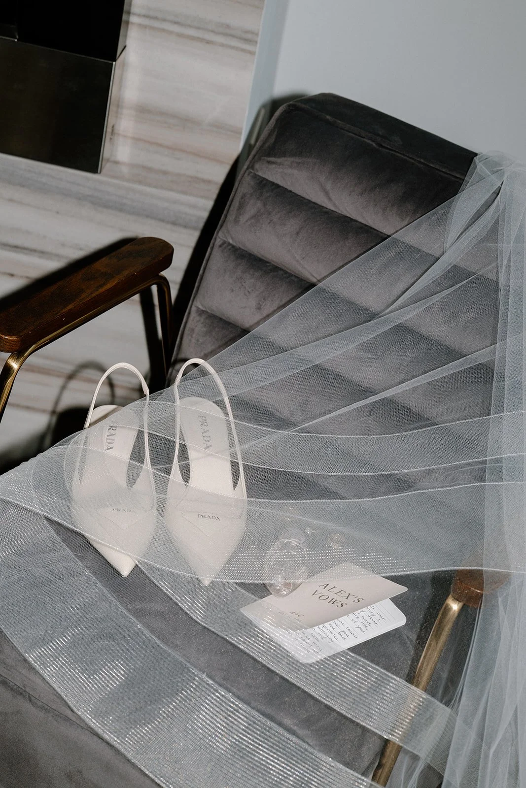 A pair of white Prada high heels on a dark gray velvet chair, with a white tulle veil draped over the chair. There is also a small card and a handwritten note on the chair.
