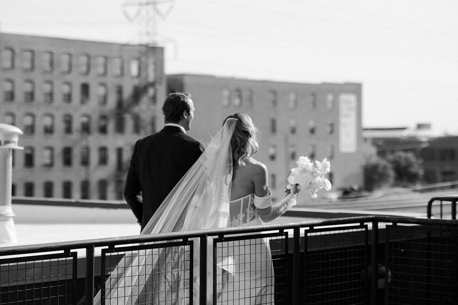 A bride and groom on a rooftop, with the bride holding a bouquet, looking away from the camera in black and white.