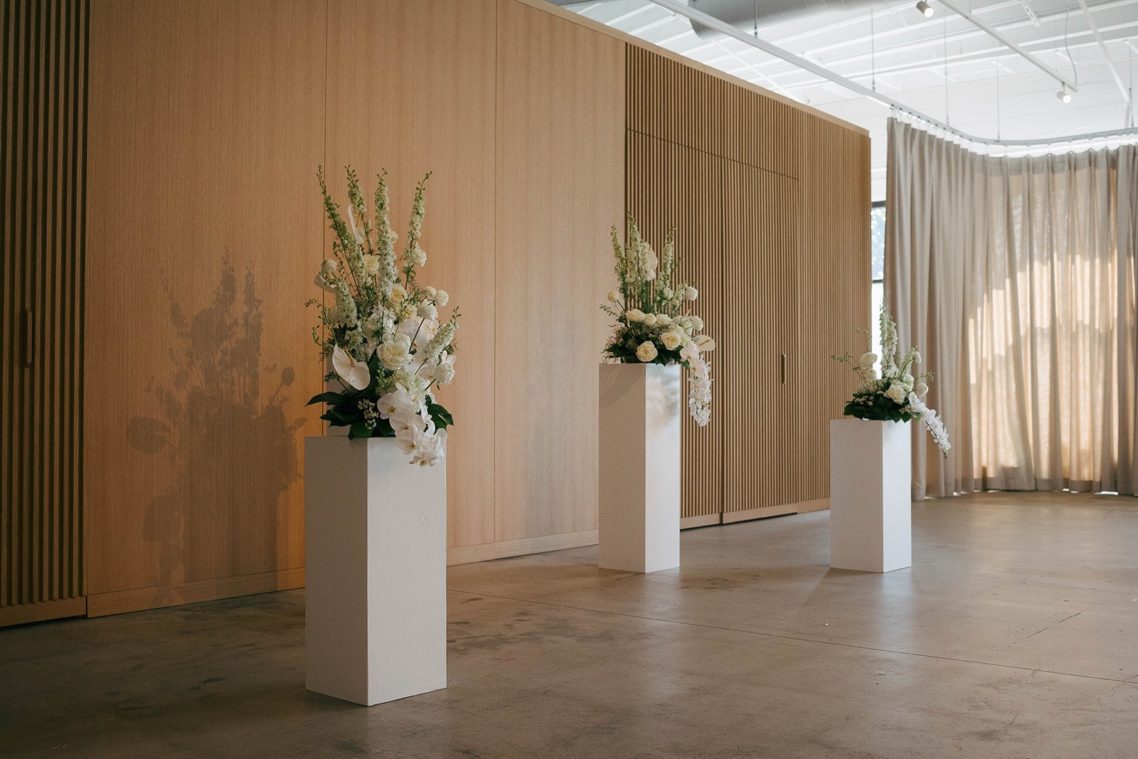 Three white pedestals with white floral arrangements in a modern, minimalistic indoor space with wooden walls and large windows with curtains.