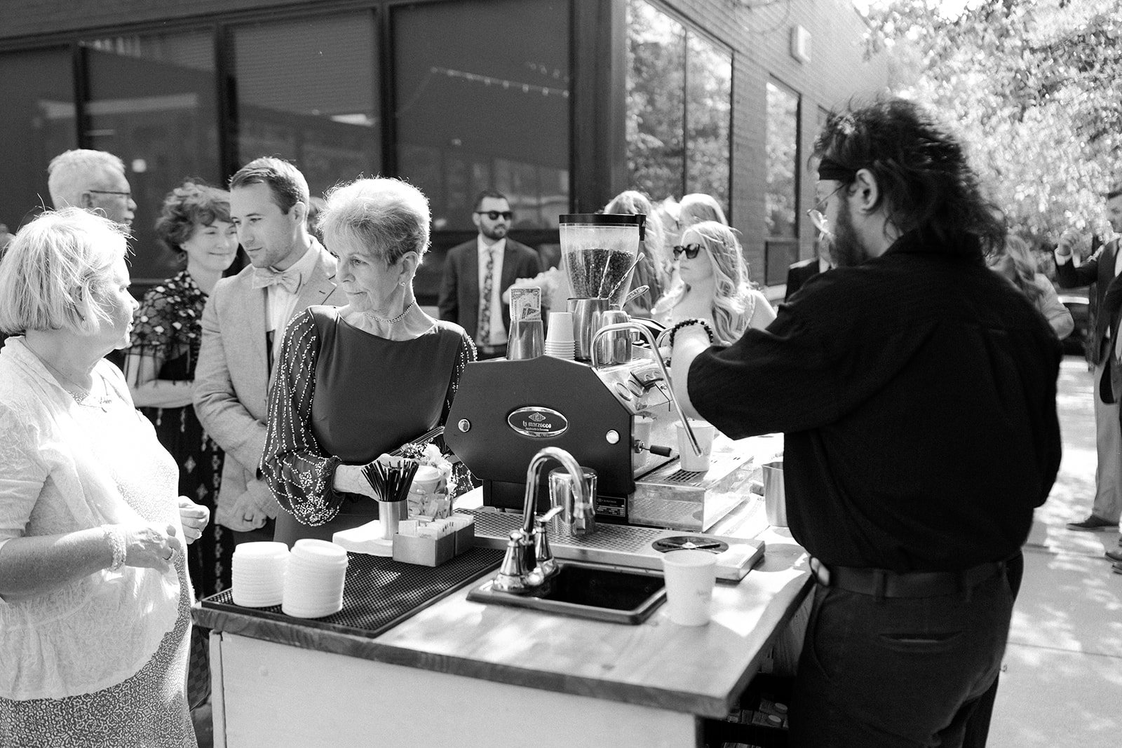 Group of people in line at a coffee stand outdoors, with the barista preparing a drink, set against a sunny day and modern building.