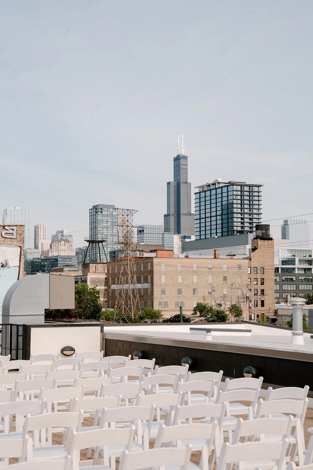City skyline with tall buildings, including the Willis Tower, seen from a rooftop with white chairs arranged in rows facing away from the camera.