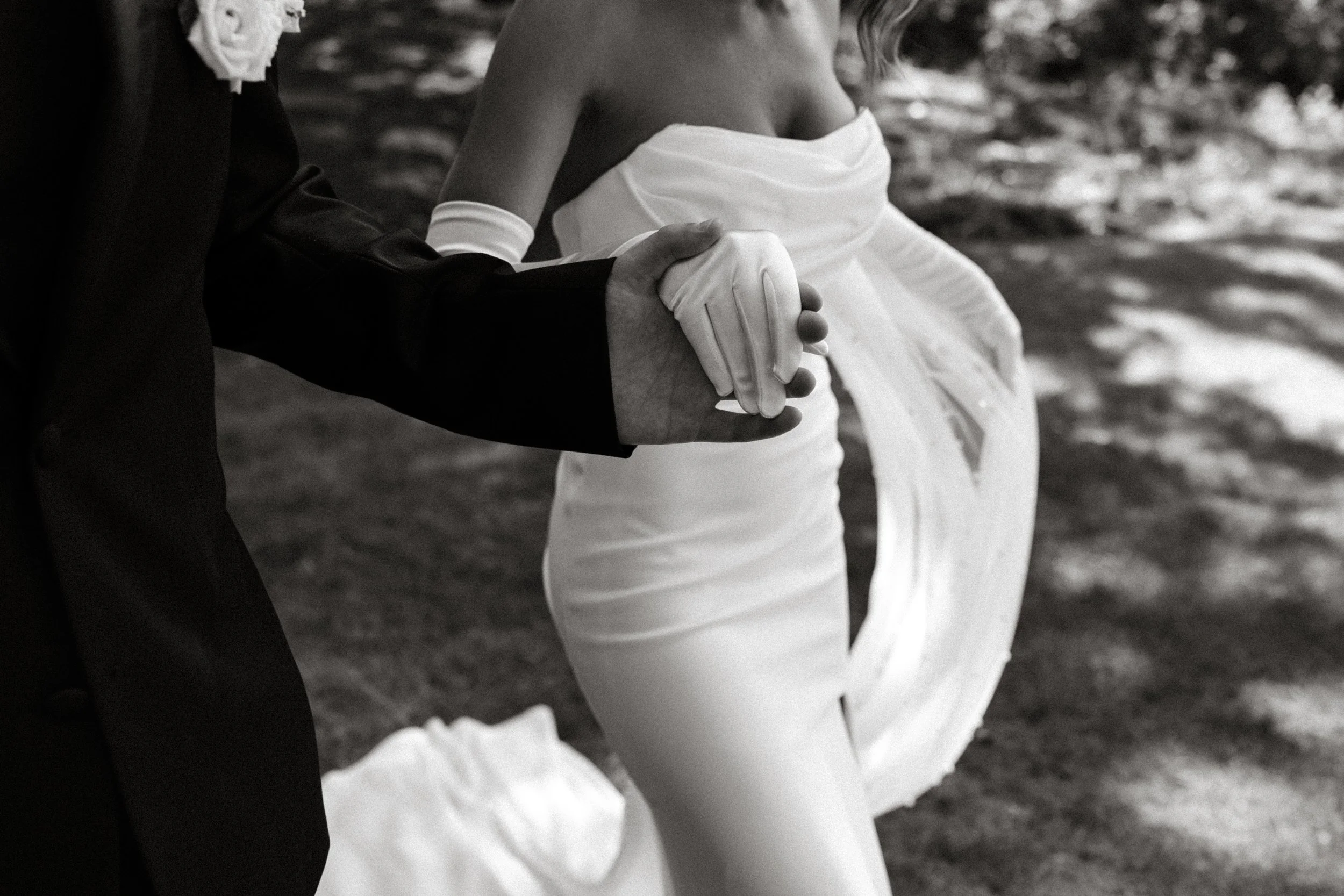 A bride and groom holding hands during their wedding. The bride is wearing a strapless wedding dress and gloves, while the groom is in a dark suit. They are outdoors, possibly during their wedding ceremony.