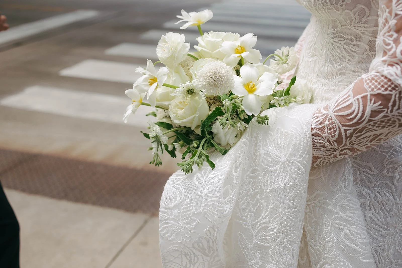 A bride holding a bouquet of white flowers, including roses and daisies, while wearing a lace wedding dress.