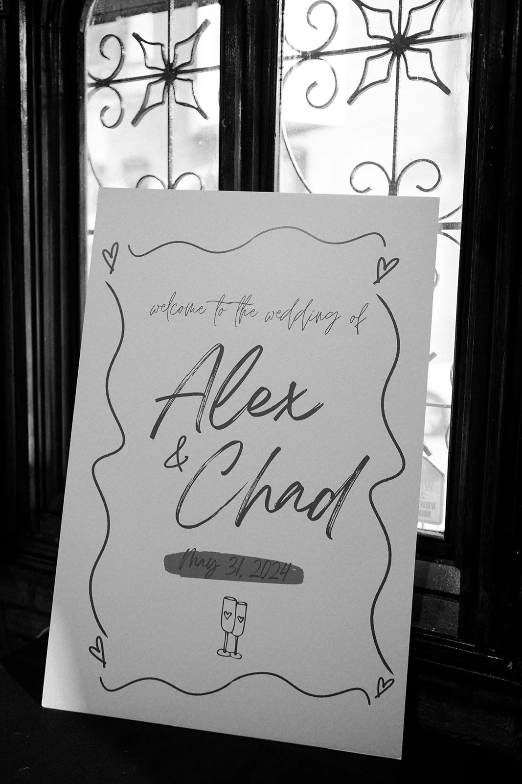 Wedding welcome sign with decorative border, featuring handwritten text that says "welcome to the wedding of Alex & Chad" with the date May 31, 2024, and a drawing of two champagne glasses with hearts.