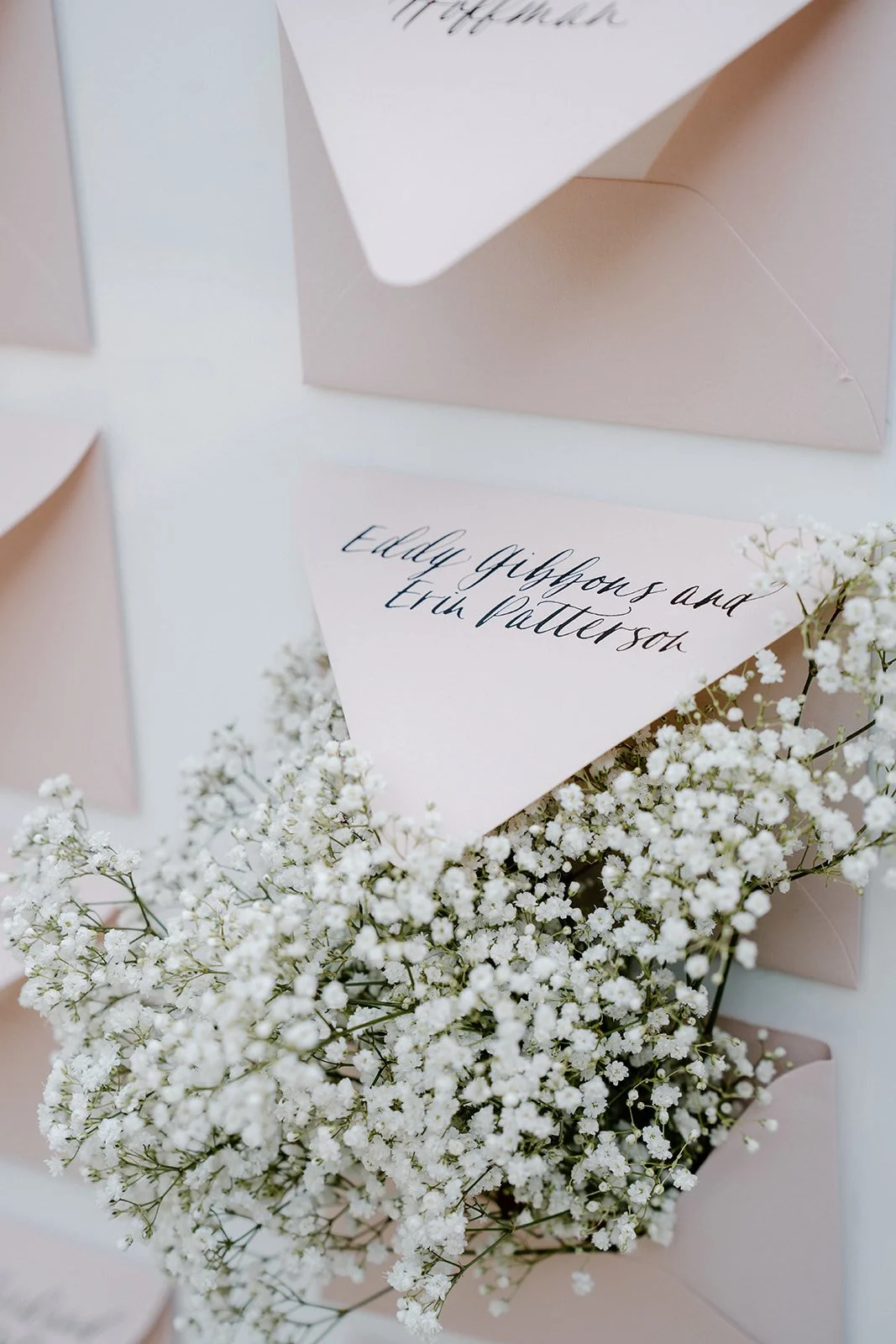 A wedding invitation with calligraphy text placed among white baby's breath flowers and surrounded by light pink envelopes.