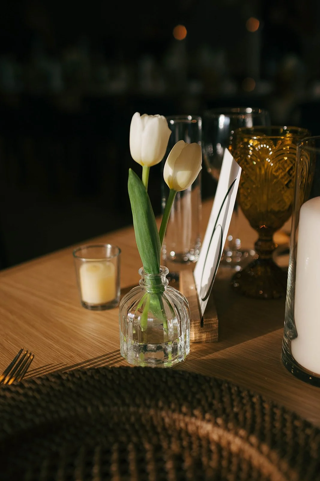 White tulips in a small clear glass vase on a wooden table, surrounded by candles and elegant glassware, with soft lighting creating a cozy ambiance.