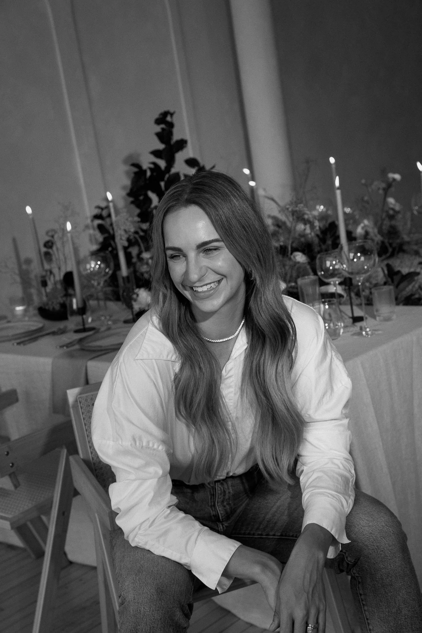 A woman with long wavy hair smiling, seated in a decorated dining area with candles and flowers in the background.