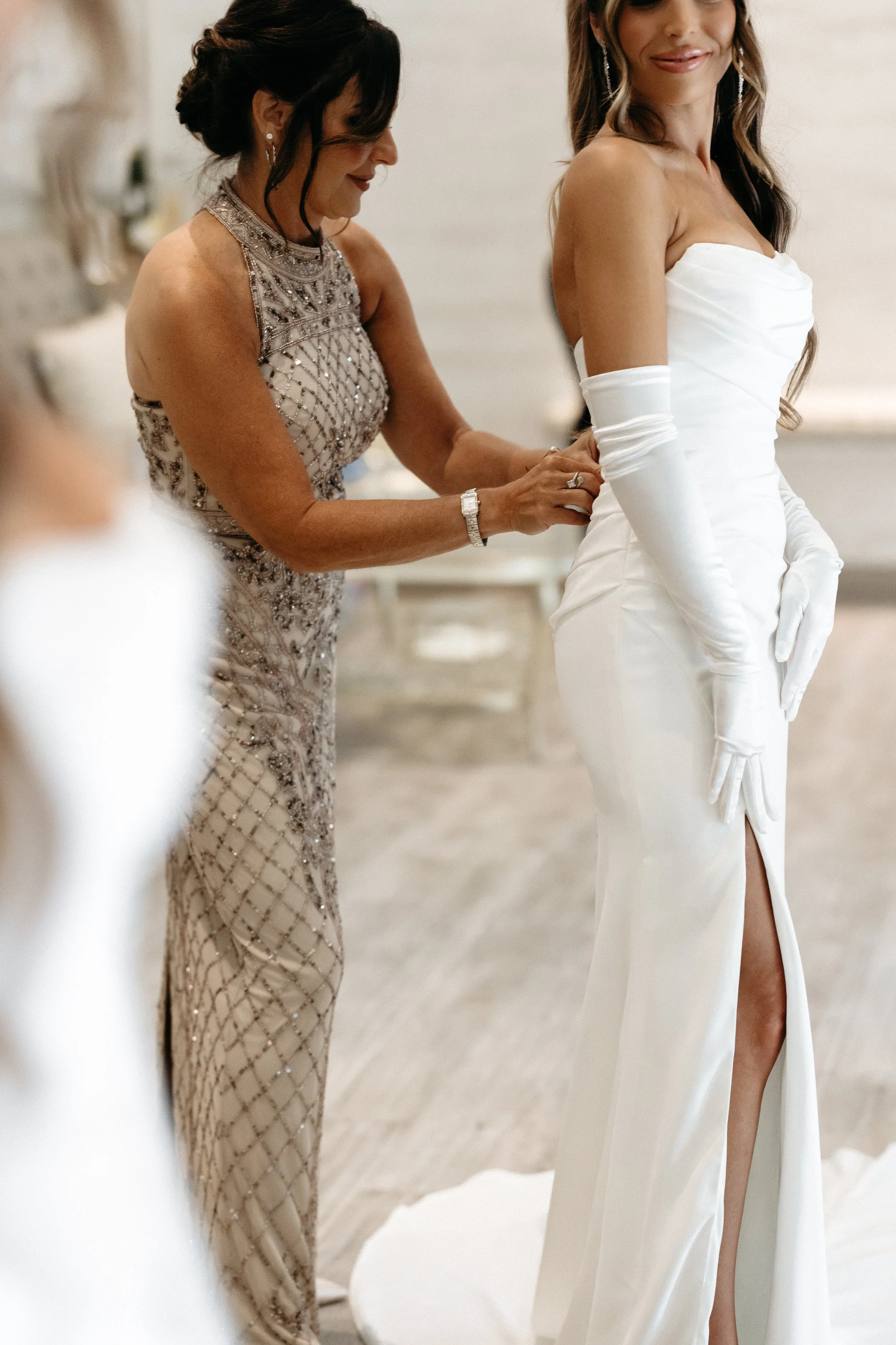 A bride in a white strapless wedding gown with long matching gloves standing in a room with a woman adjusting her dress.