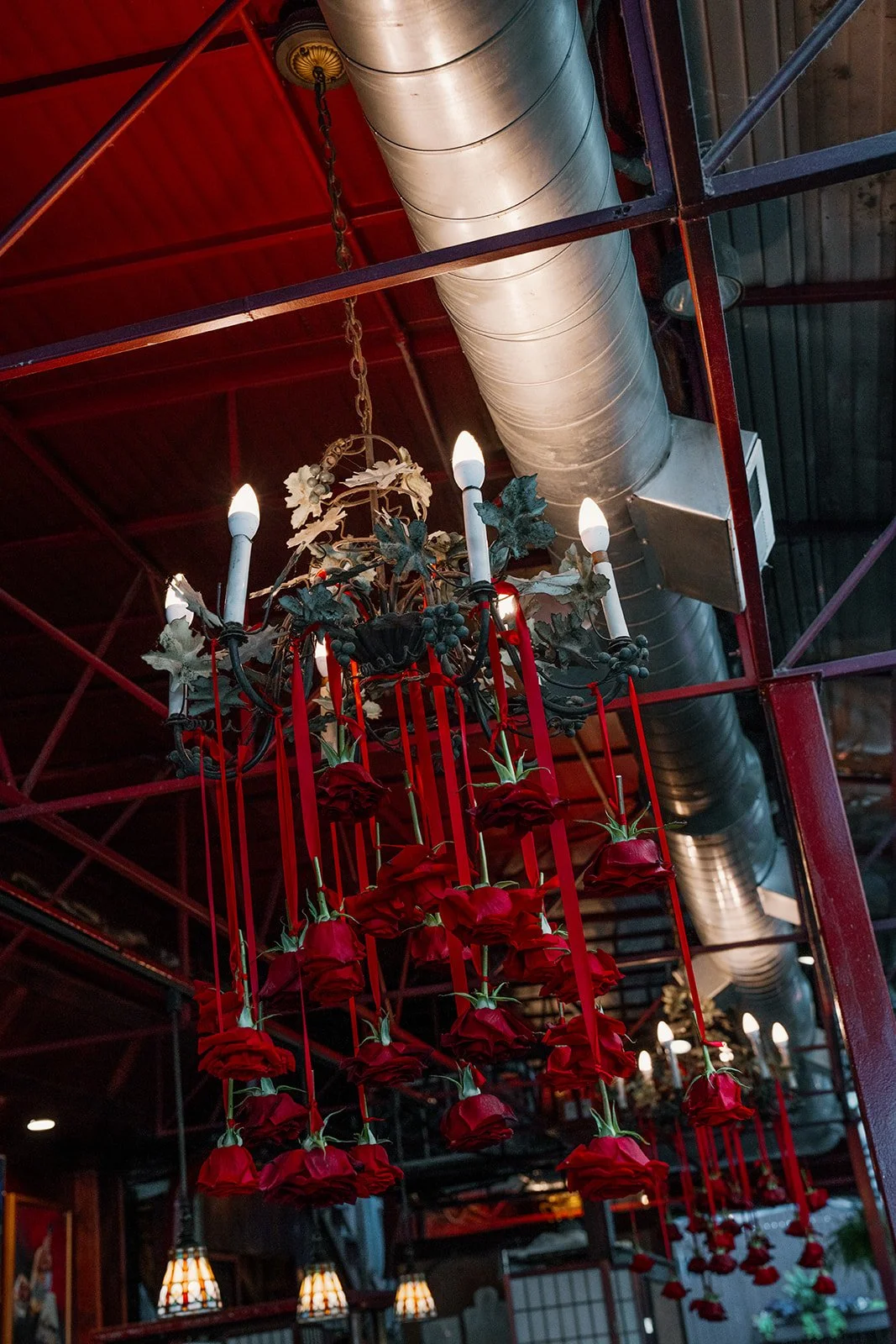 A chandelier decorated with red roses and red ribbons hanging in a restaurant or cafe with industrial ceiling and ductwork.